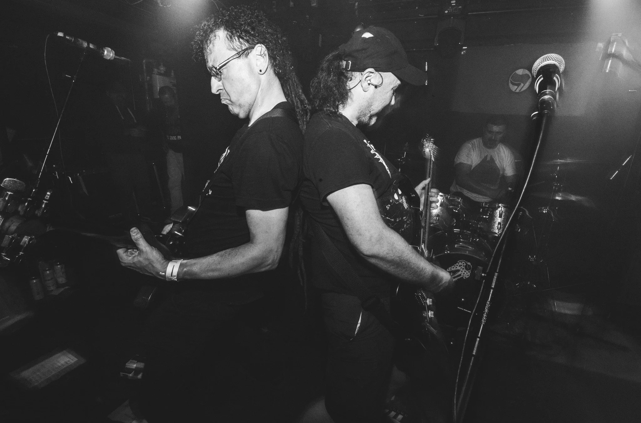 Two musicians with long hair and glasses, back to back, playing guitars on stage in a dimly lit venue.