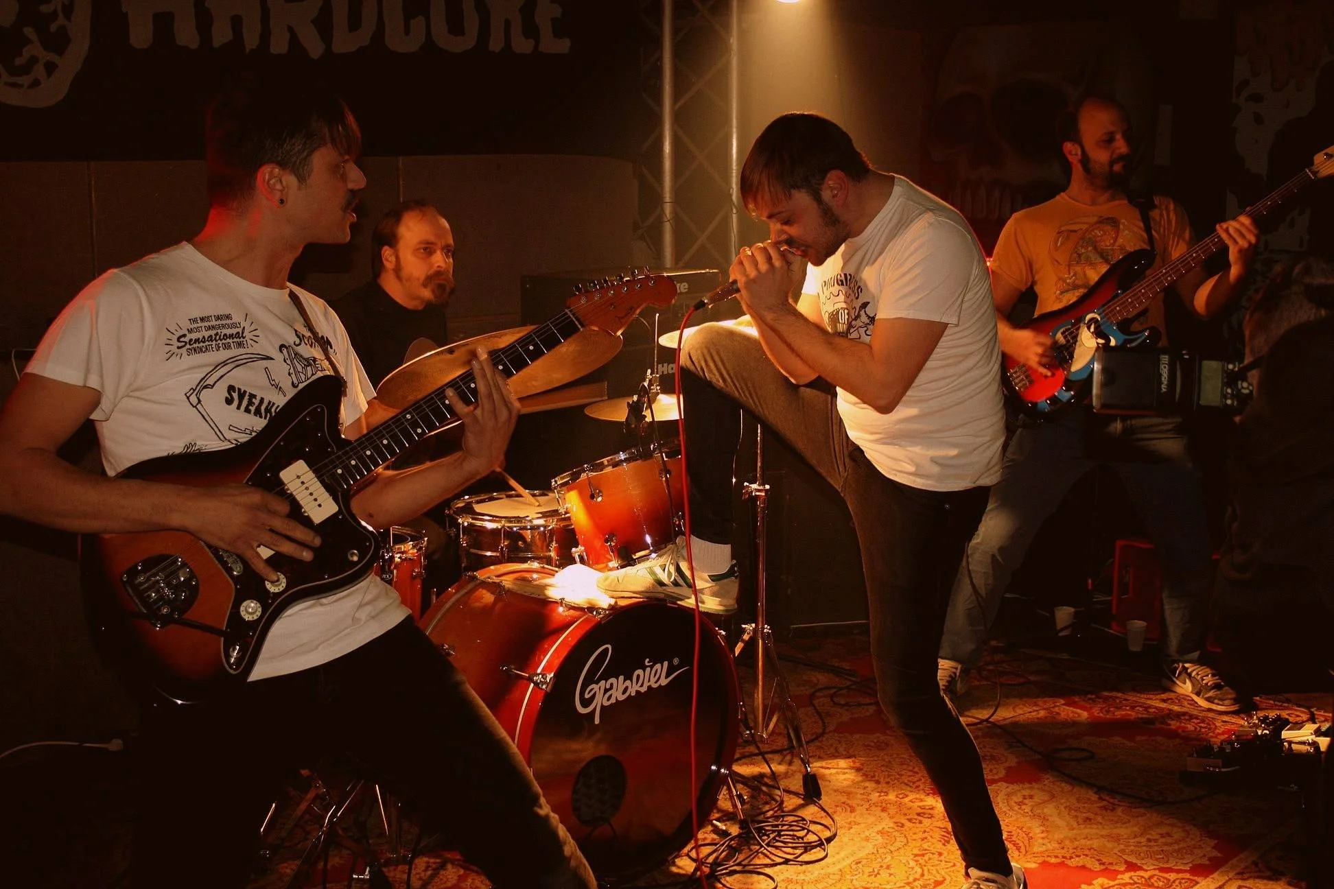 A band performing live on stage in a dimly lit venue. The scene features a guitarist playing an electric guitar, a drummer, a vocalist singing into a microphone, and bass players. The background includes a black backdrop with logo or text, and stage 
