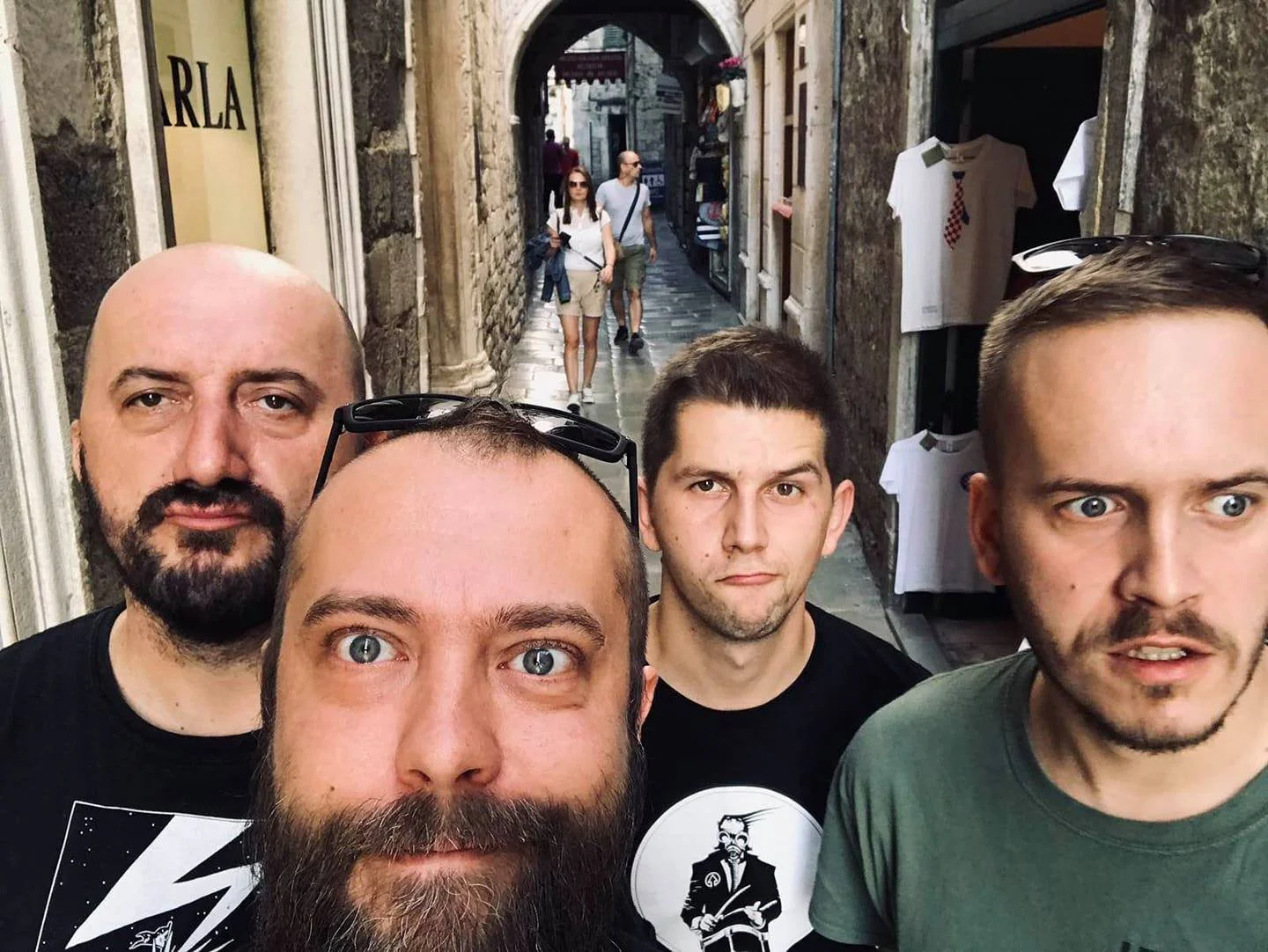 Four men taking a selfie in a narrow European street with cobblestone pavement, stone walls, shops on the sides, and pedestrians walking in the background.