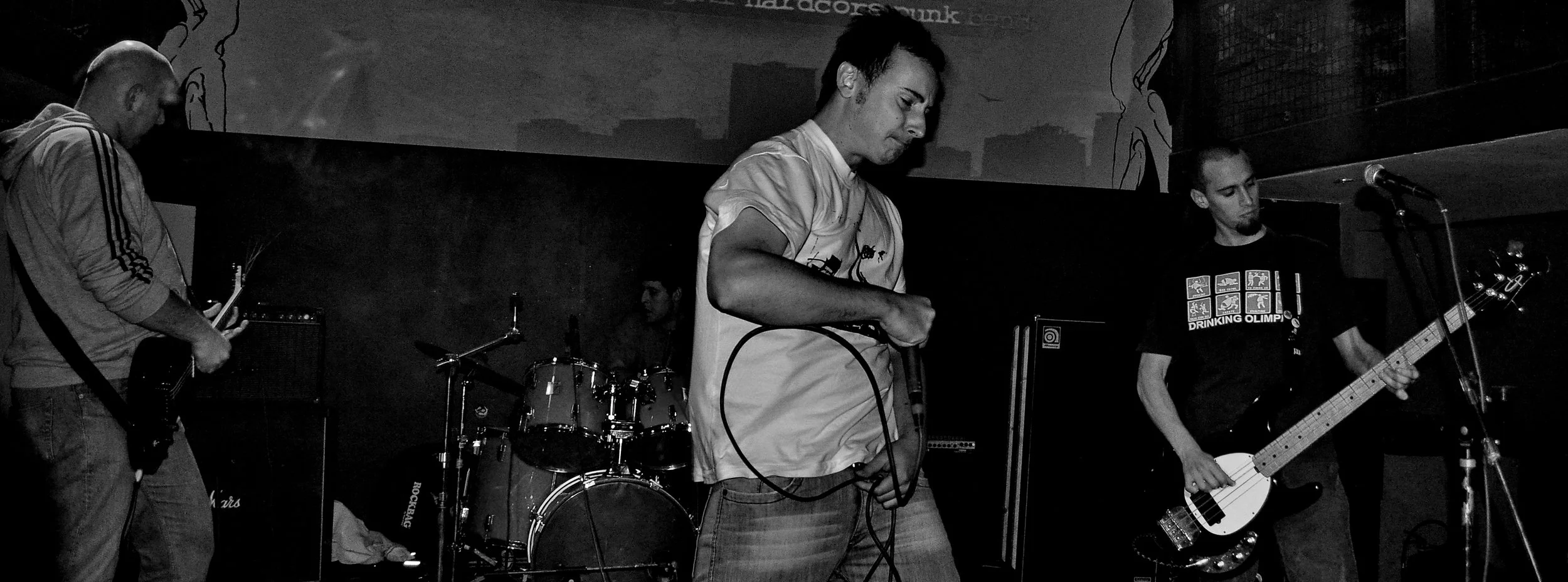A black and white photo of a band performing on stage with four members: a guitarist on the left, a vocalist holding a microphone in the center, a drummer in the back, and a bassist on the right. The stage has a dark background with some artwork and 