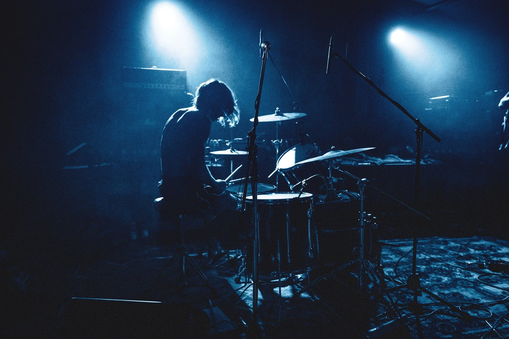 A person with long hair playing drums on stage, illuminated by blue stage lights, with microphone stands and musical equipment visible in the background.