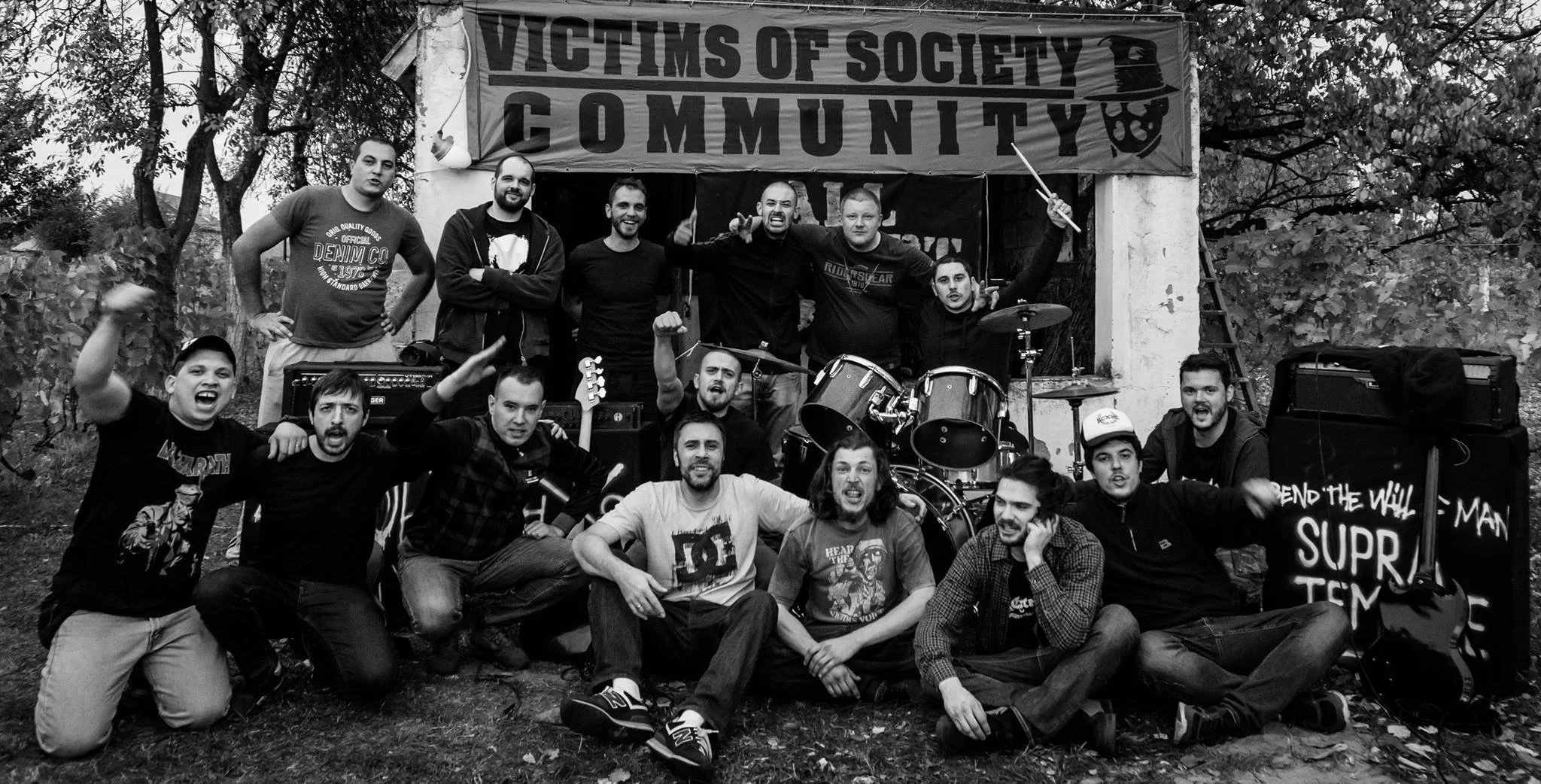A black and white photo of a group of people gathered outdoors in front of a stage with a banner that reads 'Victims of Society Community.' The group is posing for the camera, with some making gestures and smiling. There are musical instruments like 