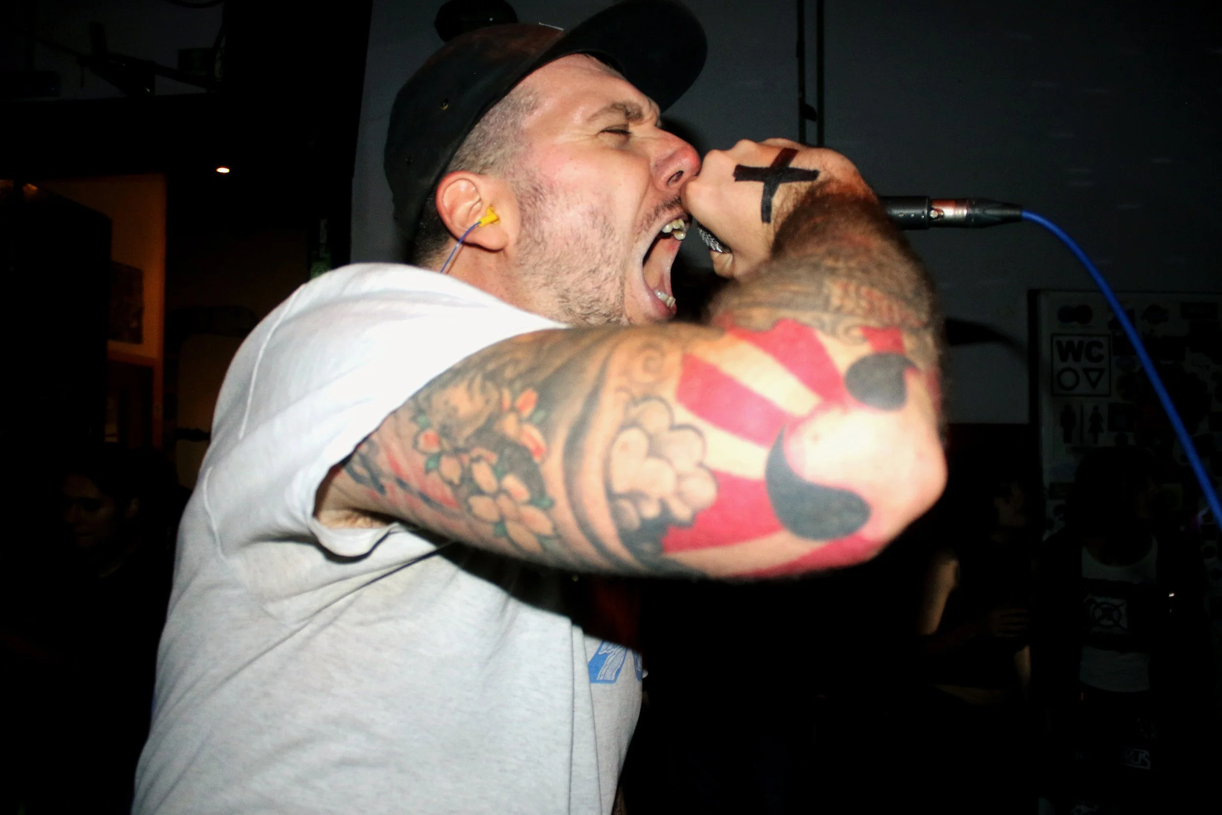 A man with tattoos on his arm and ear piercings singing or shouting into a microphone during a performance.