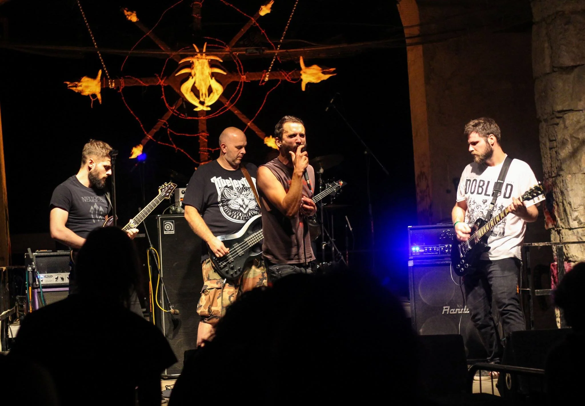 A band performing live on stage, featuring four male musicians with guitars, one singing into a microphone, and a large decorative lighting fixture with a dragon design hanging overhead.