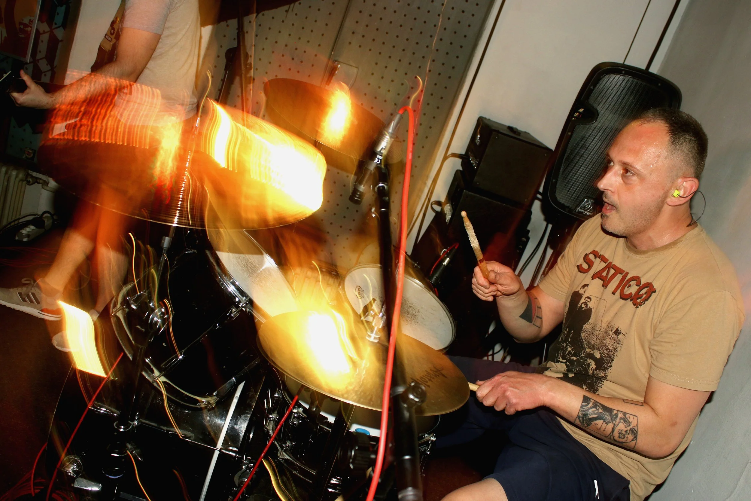 A person with tattoos on their arm, wearing a beige T-shirt with a graphic, is sitting at a drum set, holding drumsticks, in a room with sound equipment and a large speaker. The scene includes motion blur and light streaks from the drumsticks and cym