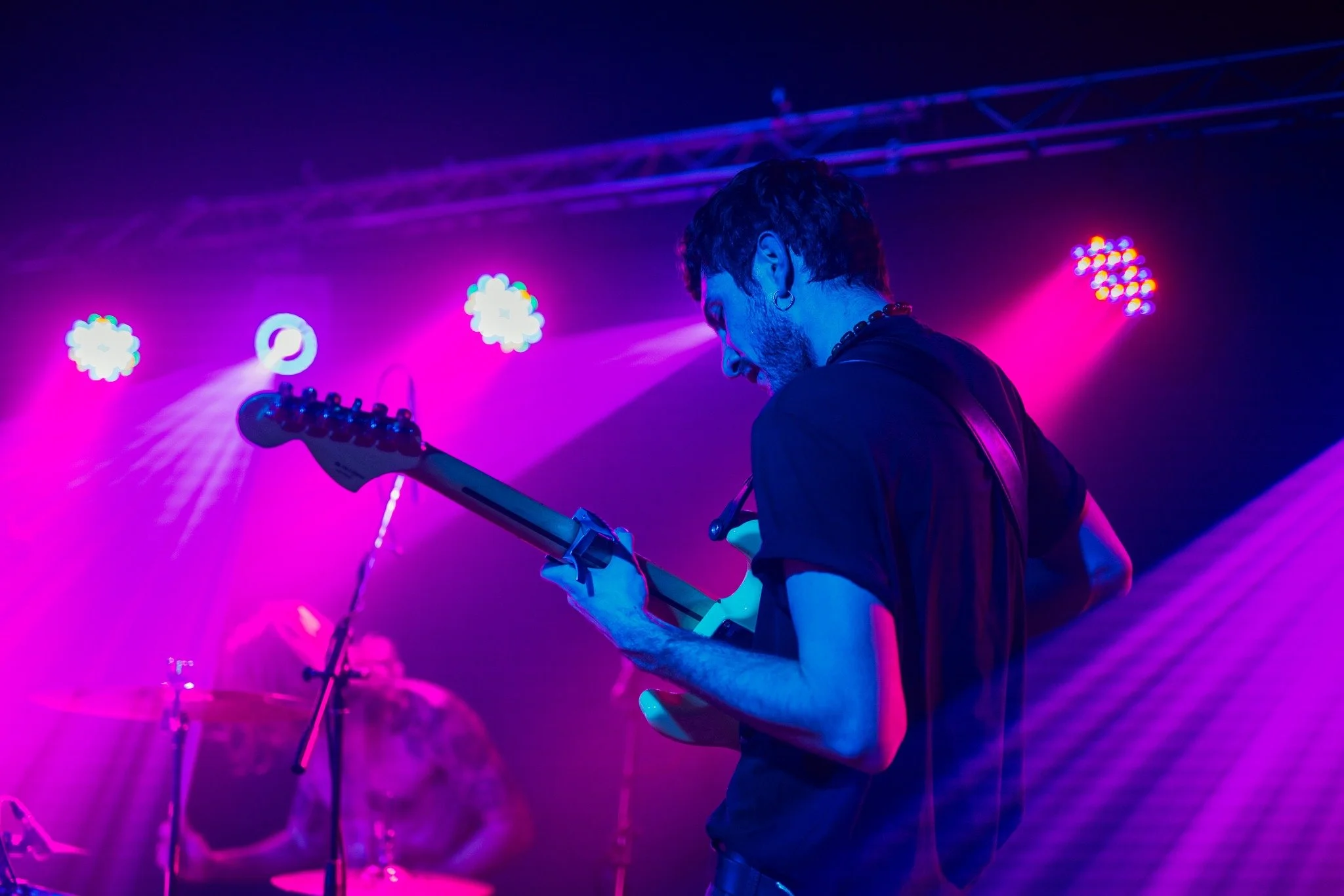 A musician playing an electric guitar on stage under colorful purple and blue stage lighting.
