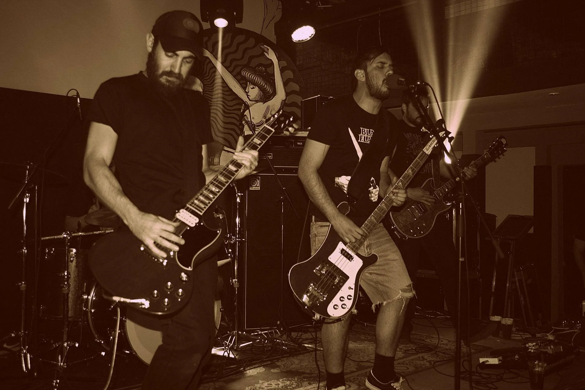 Two musicians performing on stage with electric guitars, one wearing a black t-shirt and the other in a black graphic t-shirt. The stage background includes a circular artwork featuring a woman with long hair. The scene is lit with stage lighting and