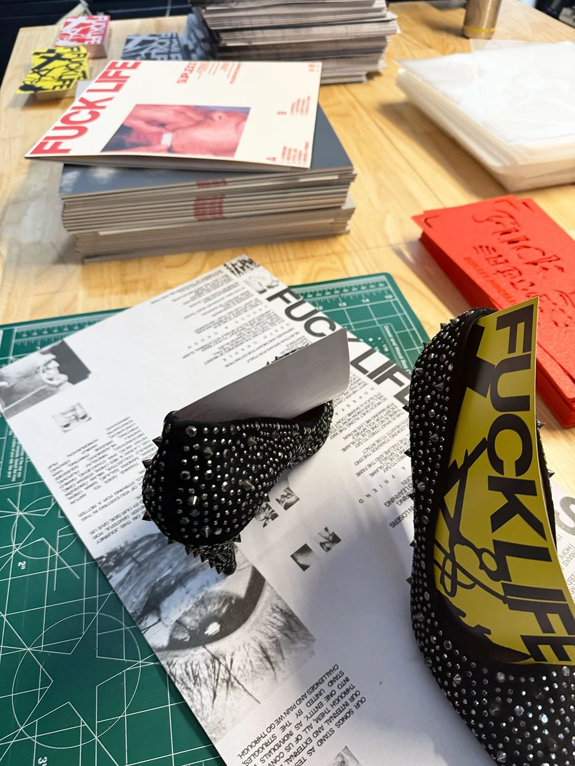 Table with stacks of magazines, comic books, and reading materials, some with bold red and yellow covers, along with black gloves covered in metal studs and a yellow and black sticker, and a cutting mat used for crafting.