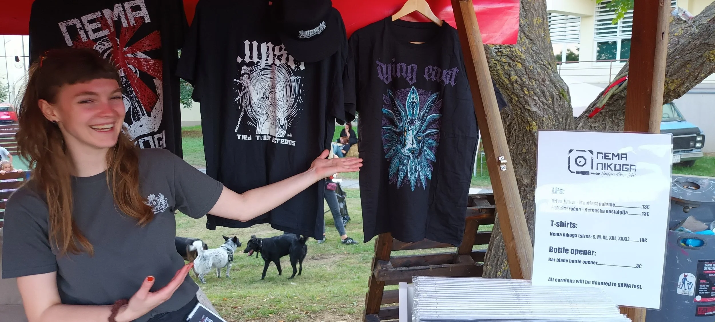 A young woman with red hair smiling at a street fair stands next to a booth displaying black T-shirts with graphic designs. There is a sign on the booth lists prices for T-shirts and other items, indicating sales benefiting a cause.