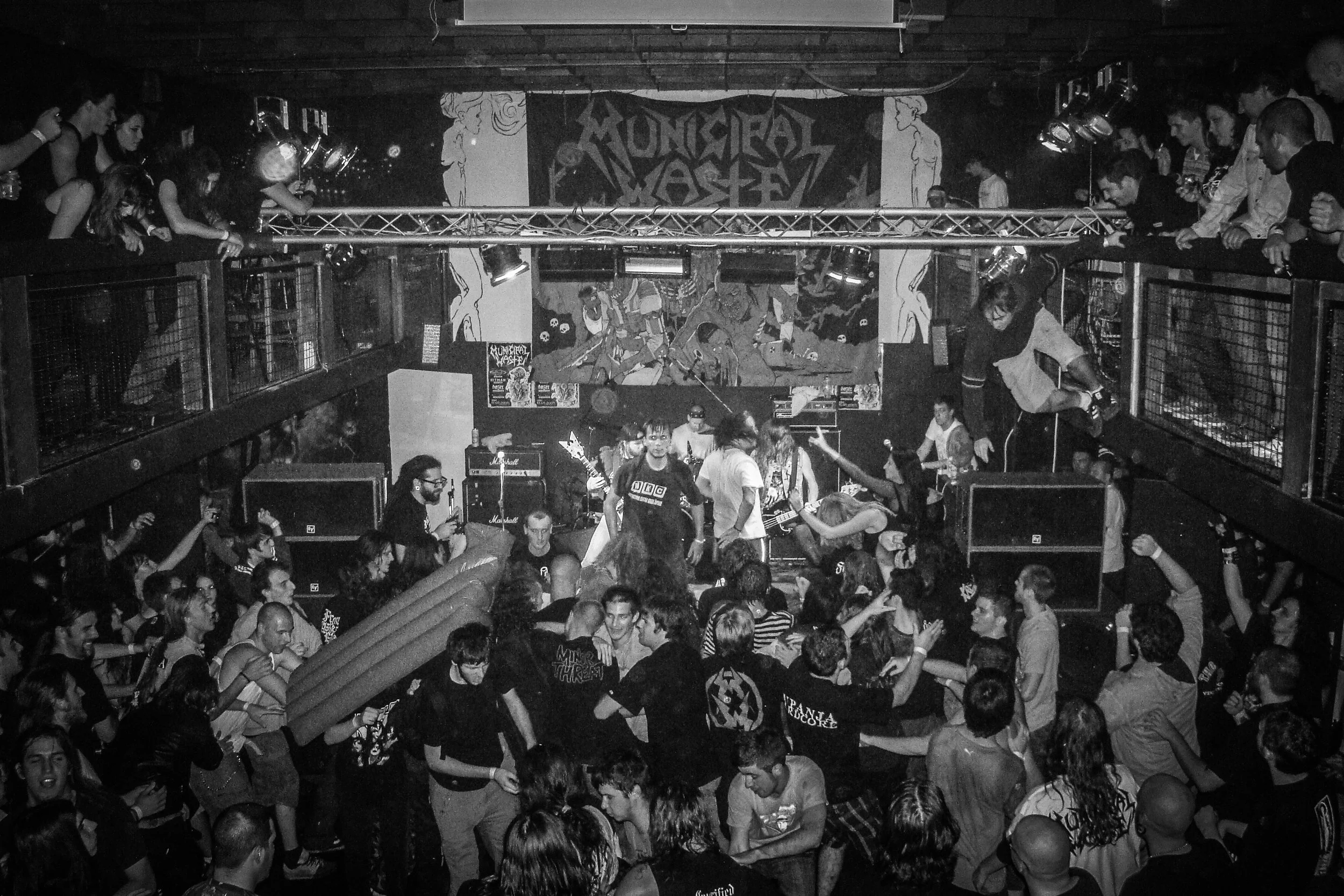 A black and white photo of a crowded indoor music venue with people standing and dancing on the main floor and balcony. There is a band playing on stage with various instruments and posters, including one that says "MUNICIPAL WASTE".