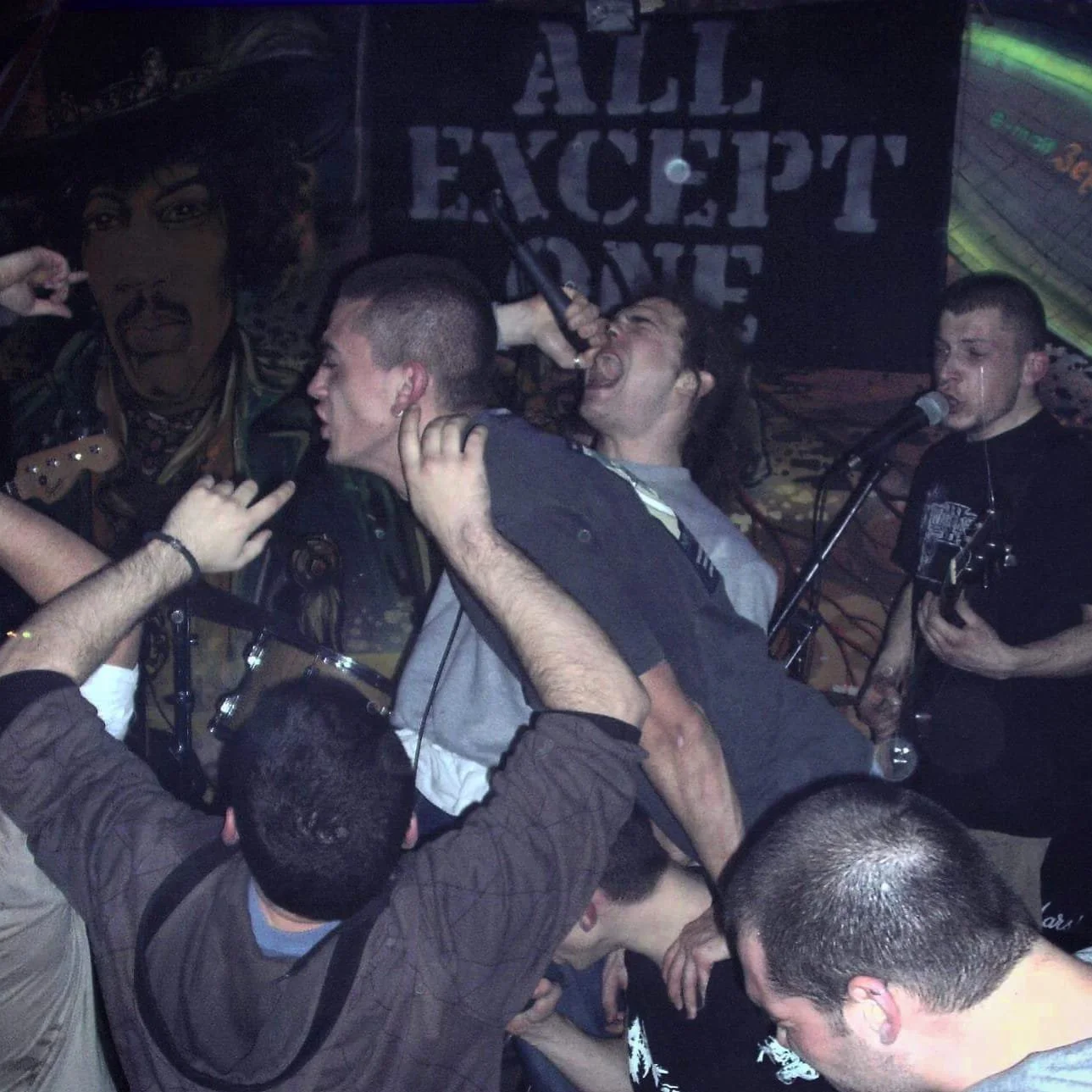 A crowded live music concert with a band on stage. A singer is passionately performing with a microphone, while the guitarist plays beside him. The background has a sign that reads 'ALL EXCEPT ONE.' Audience members are reaching out and crowding the 