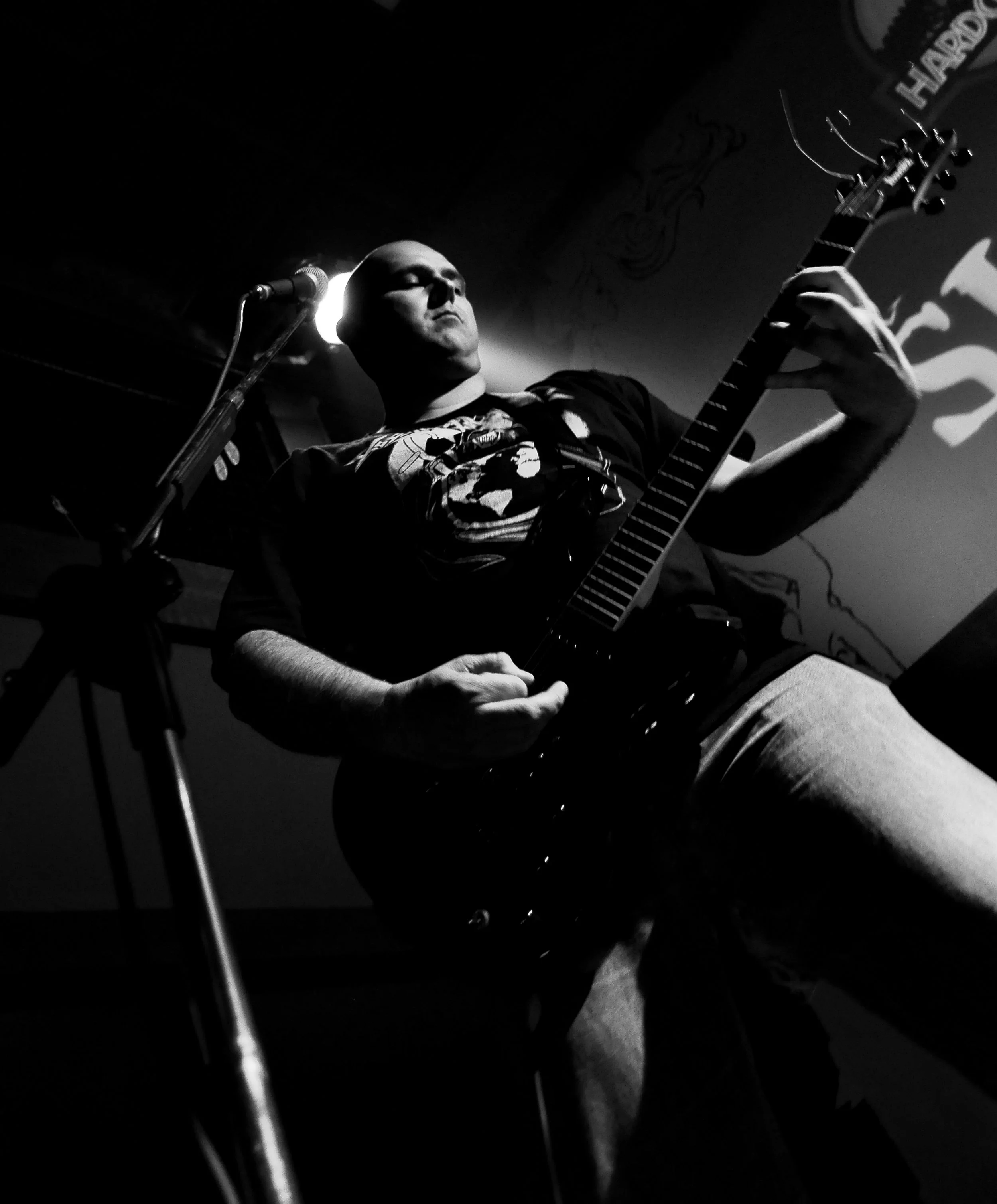 Black and white photo of a musician with eyes closed, playing an electric guitar on stage, with a microphone nearby and a tattoo visible on his arm.