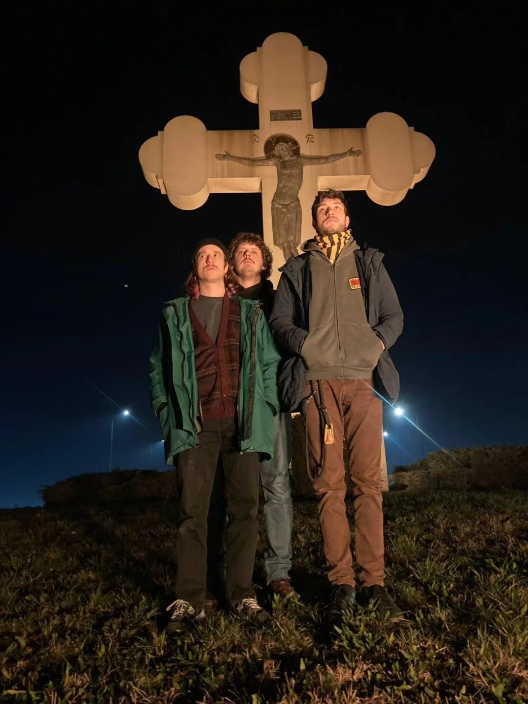 Three young men standing outdoors at night under a large illuminated cross with a depiction of Jesus Christ. All three look upward with serious expressions, dressed in casual outdoor clothing.