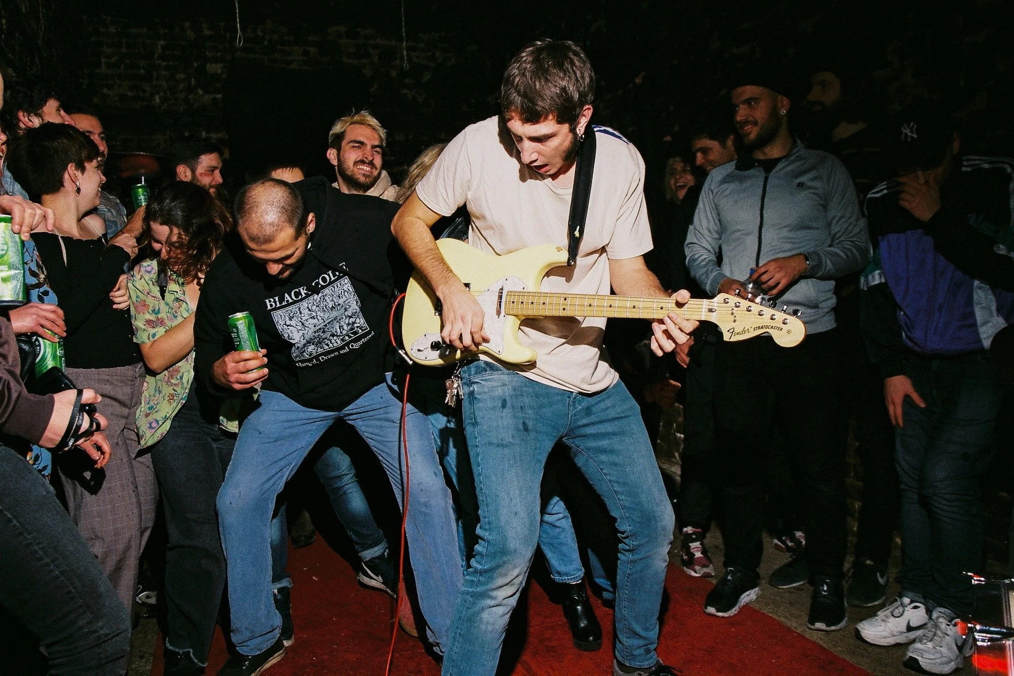 A group of people at a concert or party, with a person in the center playing an electric guitar surrounded by a crowd, some holding drinks, in a dark indoor setting.