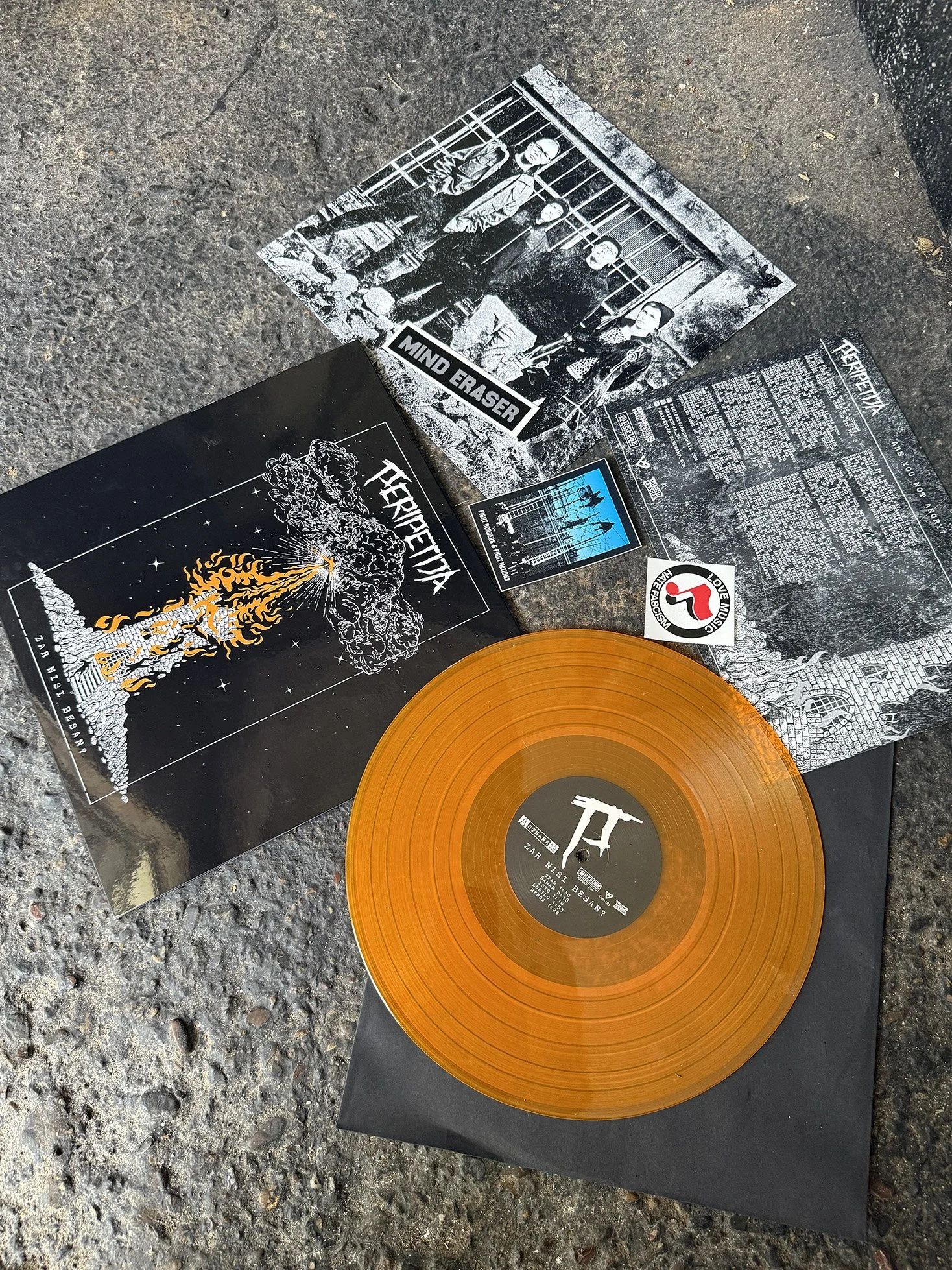 An orange vinyl record on a black sleeve, next to a black and white concert poster with a lion illustration and the band name 'TERRIFIERX', and several flyers and photos scattered on a rough asphalt surface.