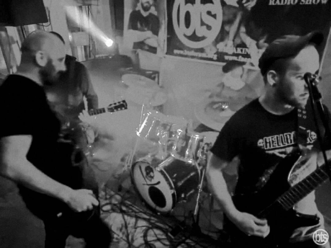 Four musicians performing live in a small venue, with a drum set, two guitarists, and a bassist, and promotional posters on the wall.