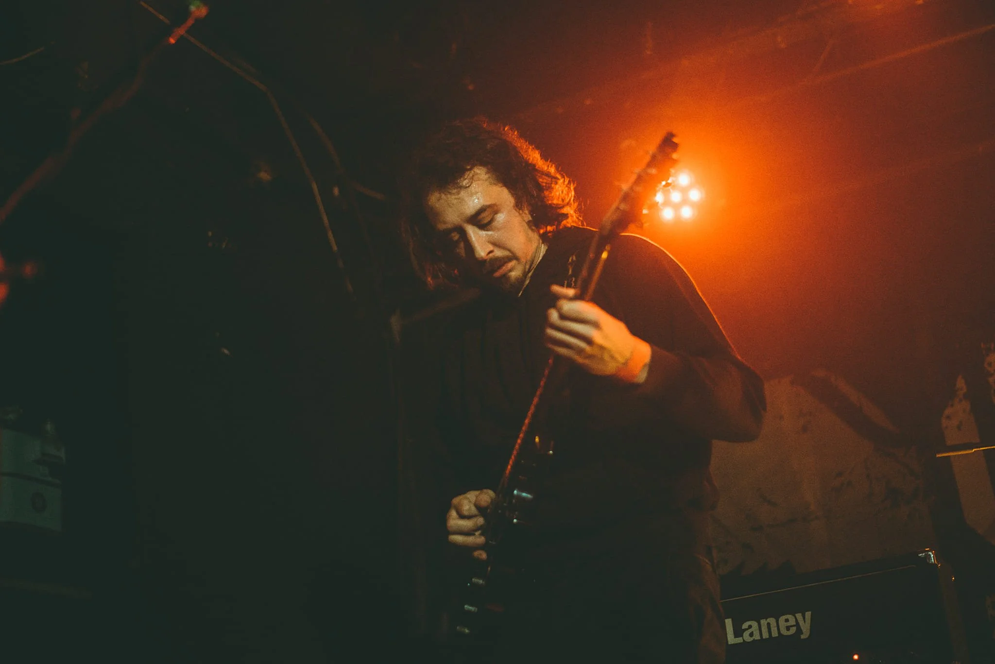 A person with long curly hair playing an electric guitar on stage with orange lighting behind them.