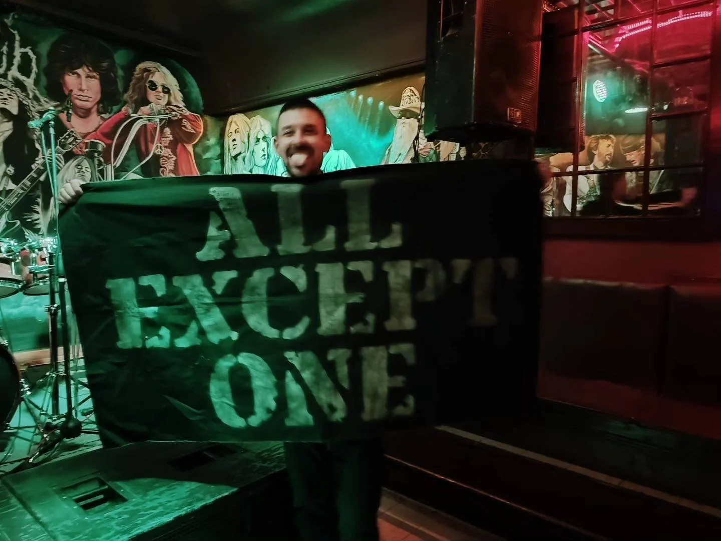 Person smiling and sticking out tongue behind a banner that says 'Tickets One' in a dimly lit bar or club with musical artwork on the wall.