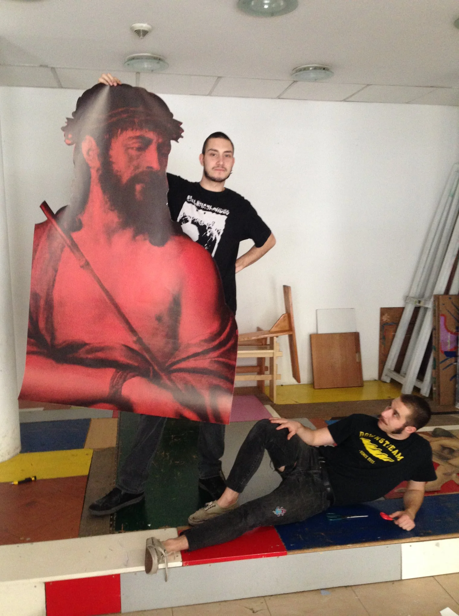 Two young men in a gym, posing with a large cardboard cutout of a bearded man with dreadlocks wearing a red shirt. One man stands behind the cutout with one hand on top, while the other man is lying on the floor, reaching out with his hand.