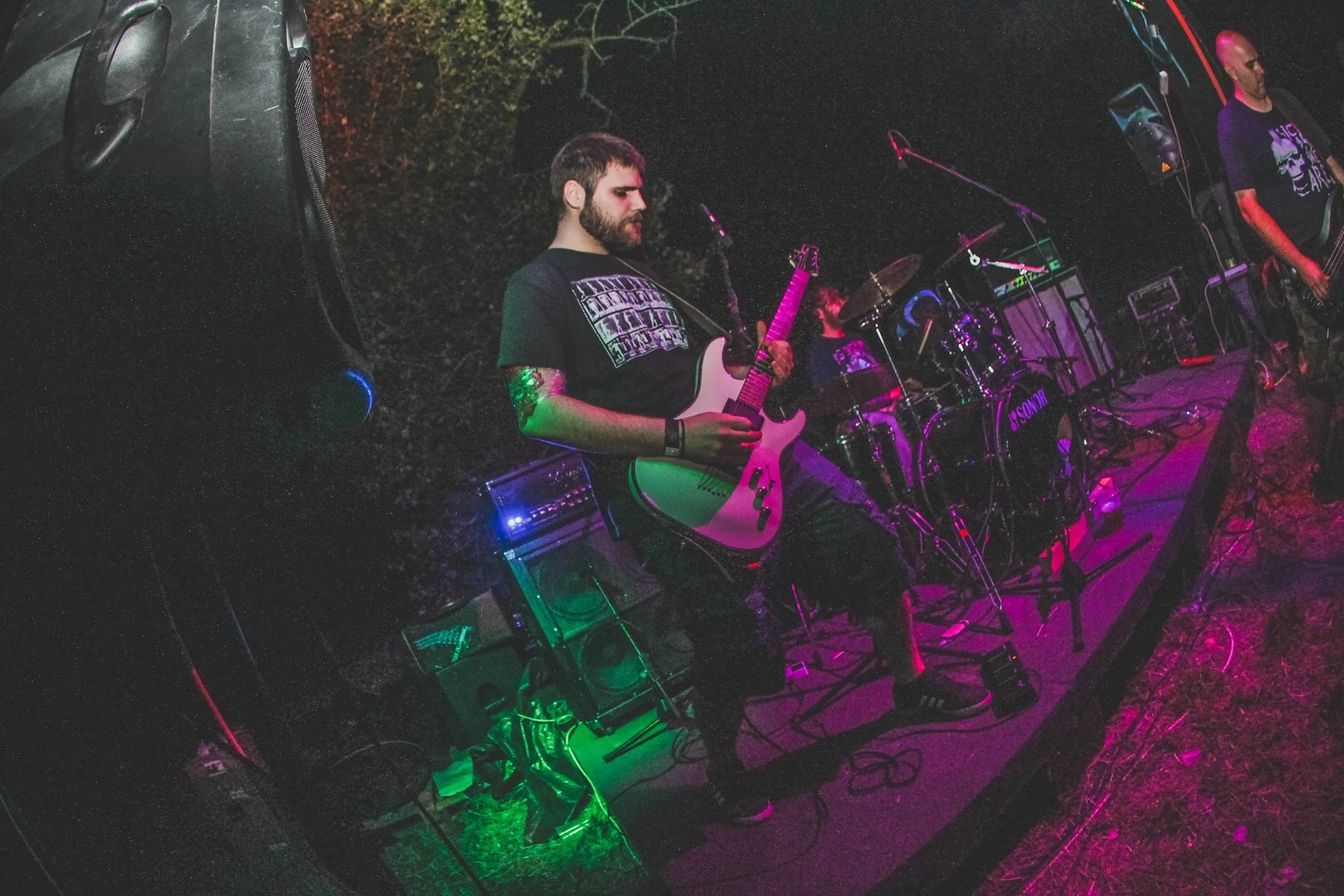 A band performing live outdoors at night with three musicians visible. One plays an electric guitar, another musician is on drums in the background, and a third person is on bass guitar. There are purple and green stage lights illuminating the scene.