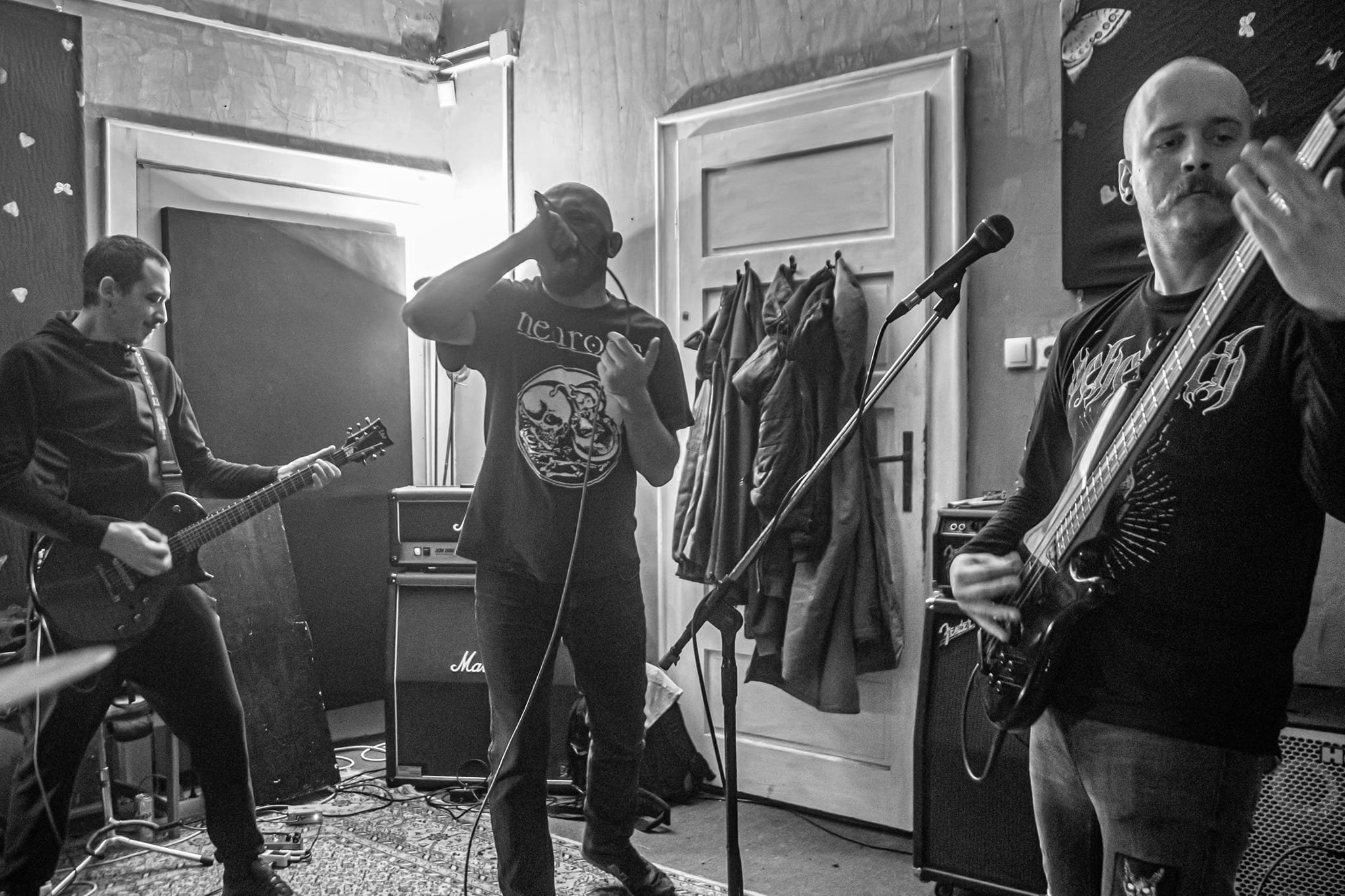 Three musicians performing in a room. One playing guitar, one singing into a microphone, and one playing bass guitar.