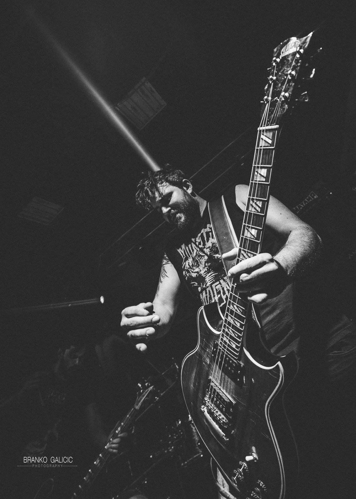 A man playing an electric guitar on stage in black and white, wearing a Metallica T-shirt, with a beard and curly hair, smiling.