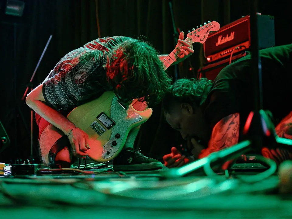Two musicians are performing on stage, one is playing an electric guitar and the other is kneeling on the floor, possibly singing into a microphone. Stage equipment and amplifiers are visible in the background.