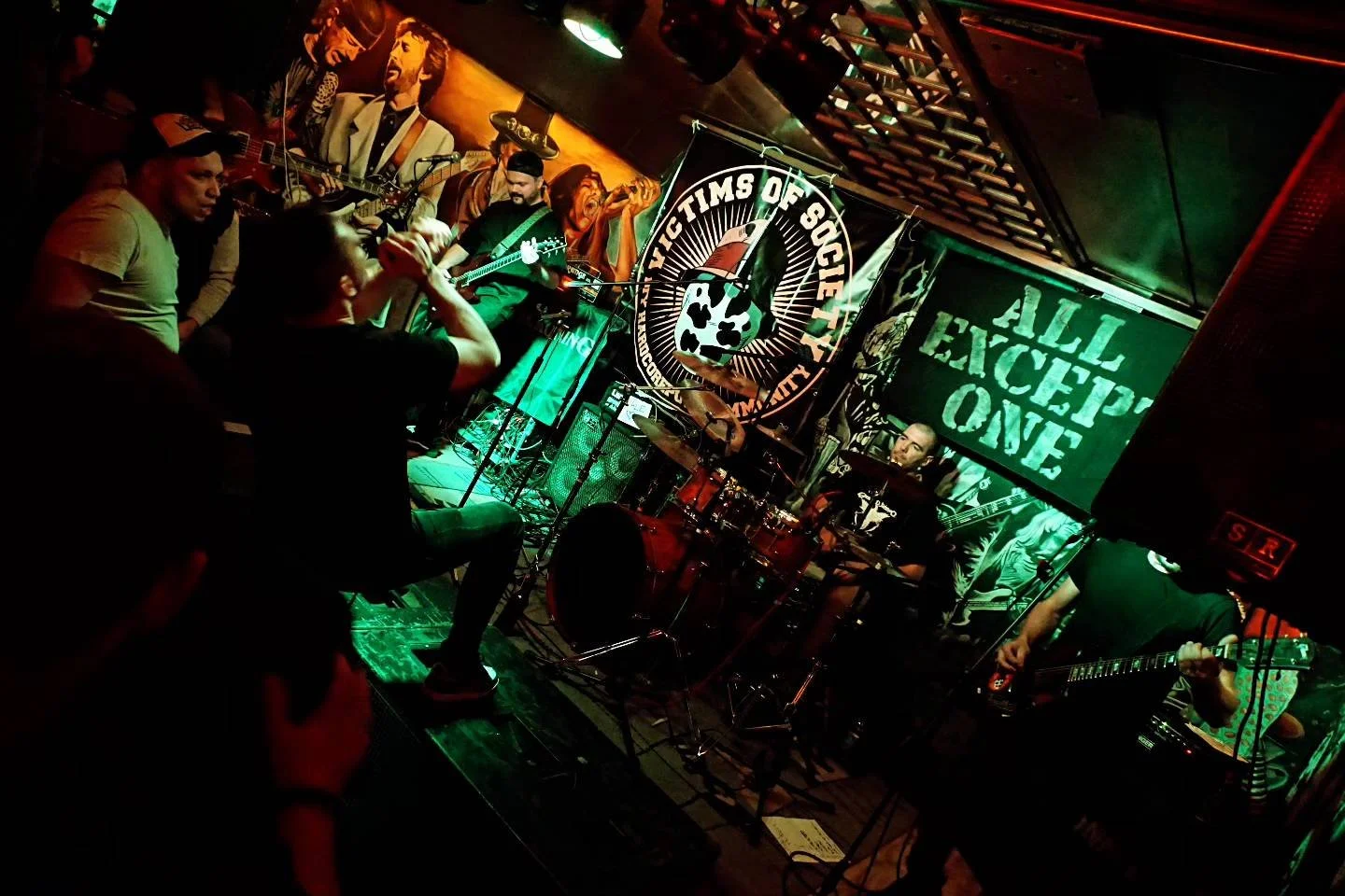 Live band performing on stage at a venue with audience watching, banners with 'Fighting in Solidarity' and 'All Except One,' murals on walls.
