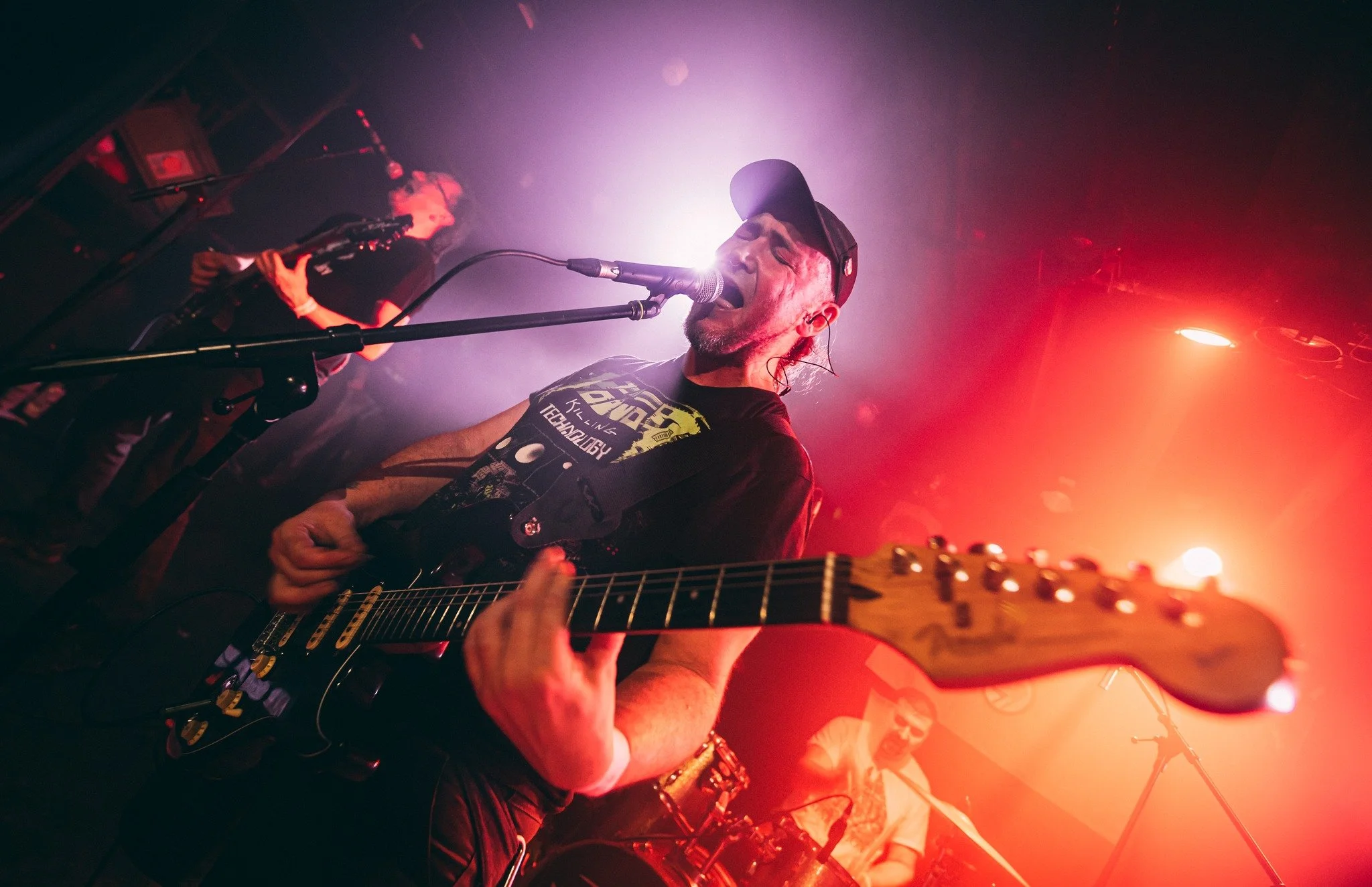 A musician wearing a black cap and Michigan Wolverines shirt plays an electric guitar and sings into a microphone during a live performance on stage with red and purple lighting.