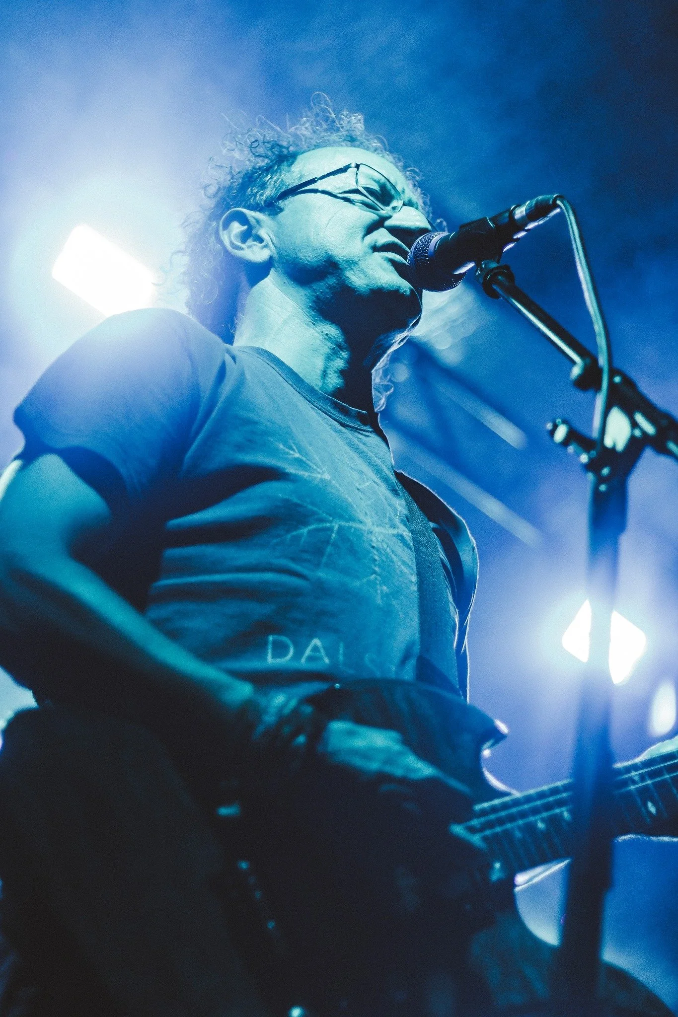 A man with curly hair, glasses, and a beard, is singing or speaking into a microphone on stage while playing a guitar.
