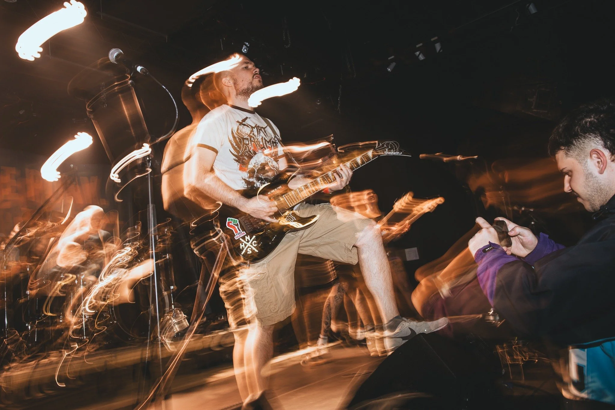 A musician playing an electric guitar on stage at a concert, with motion blur and light trails around him indicating energetic movement; a person is visible in the foreground, engaging with the performance.