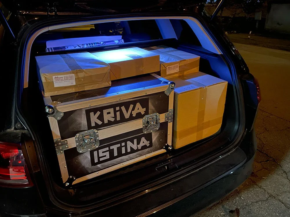 Car trunk filled with packages and a large Kriva Istina box, illuminated by interior light at night.