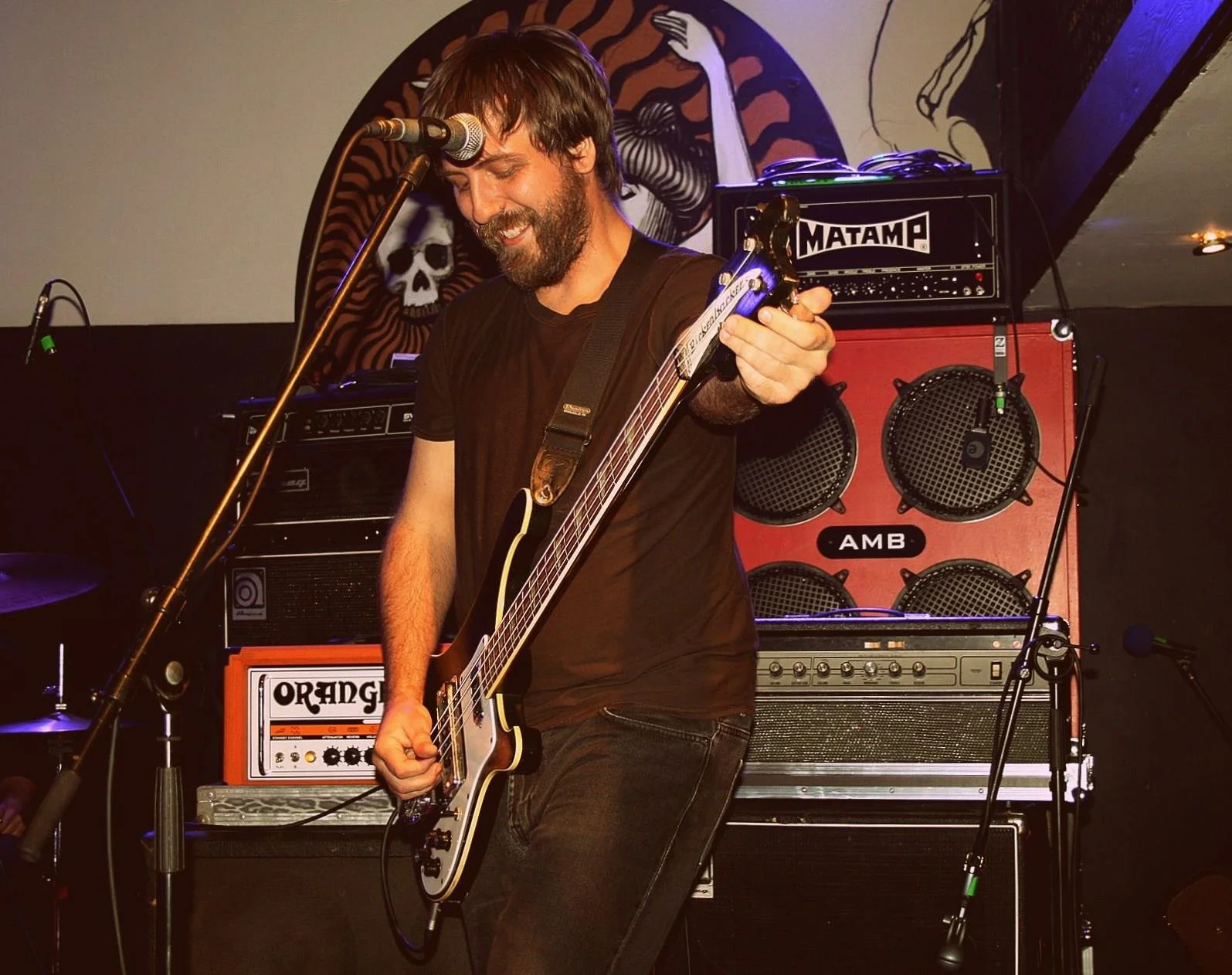 A man with dark hair and a beard, wearing a black t-shirt, playing an electric guitar on stage. Behind him, there are amplifiers and speakers, including brands Orange and Matamp. The background features a mural of a skull and a woman with flowing hai