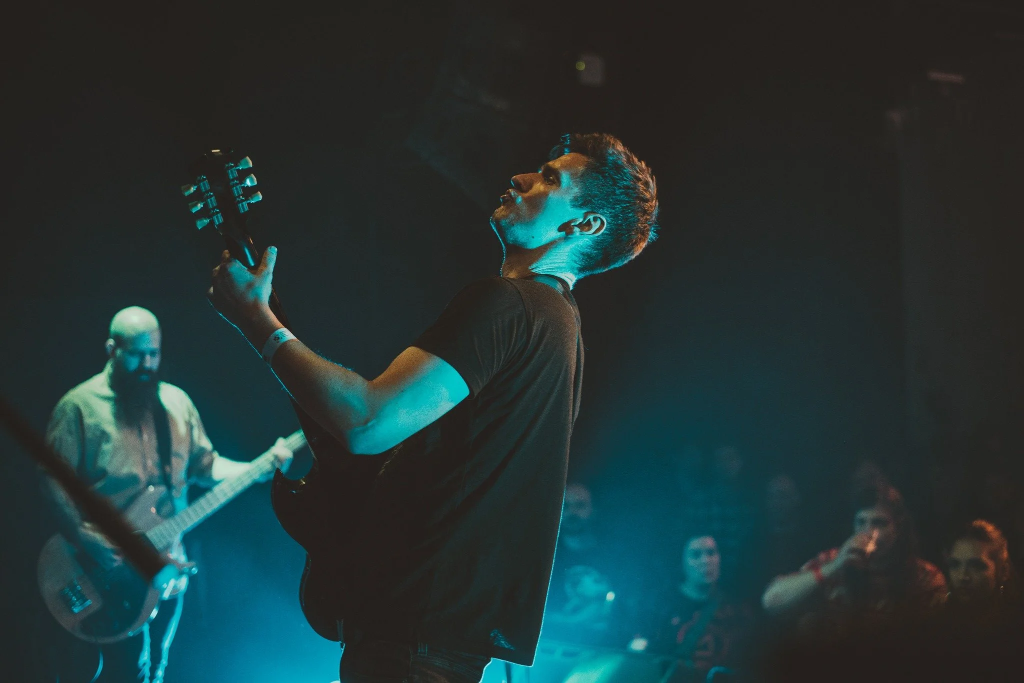 A young man playing an electric guitar on stage during a concert, illuminated by blue and teal stage lighting, with other musicians and audience members visible in the background.
