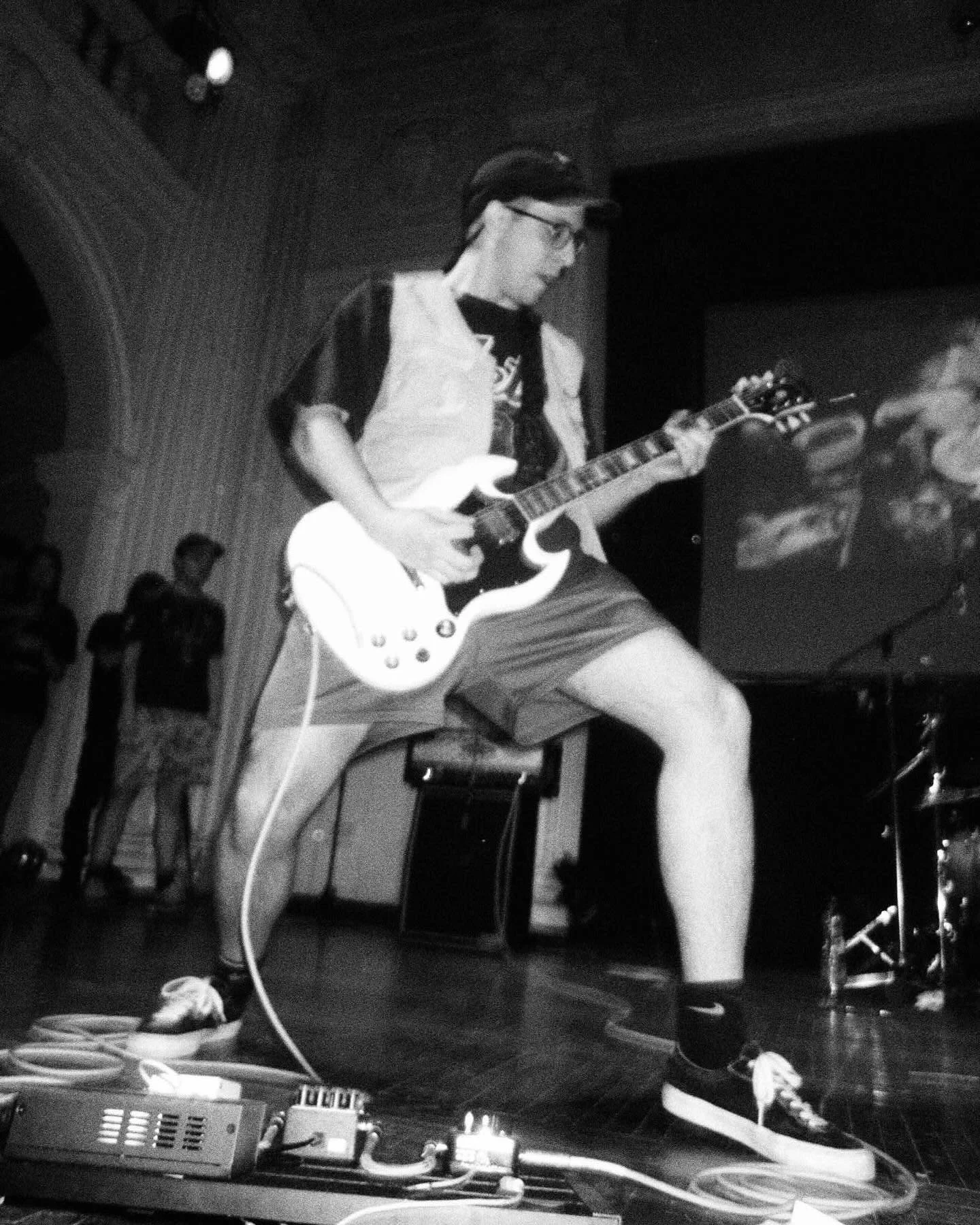 Person playing electric guitar on stage with equipment on the floor, in a dimly lit room