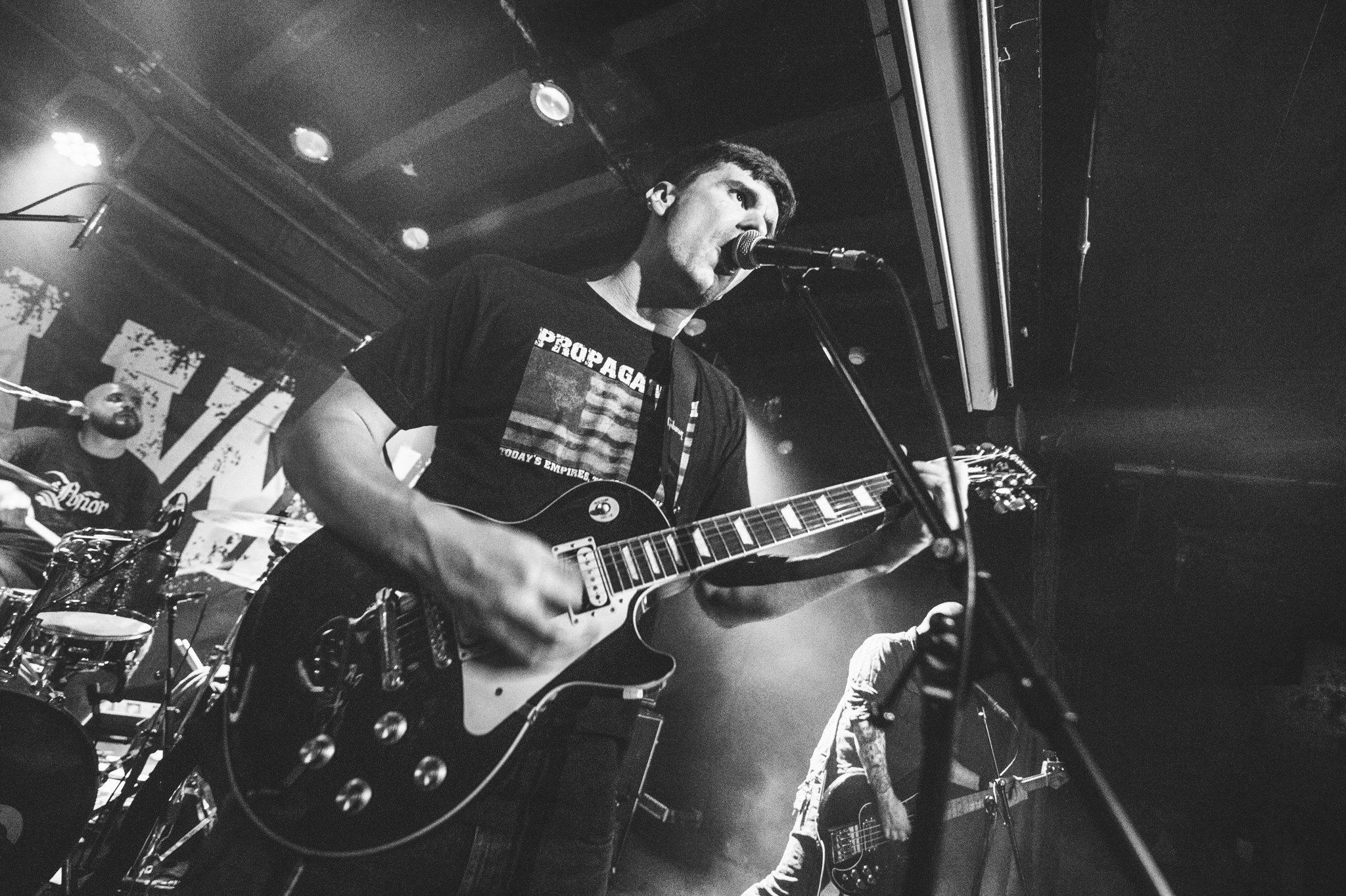 Musician singing into a microphone and playing an electric guitar on stage in a black and white photo.