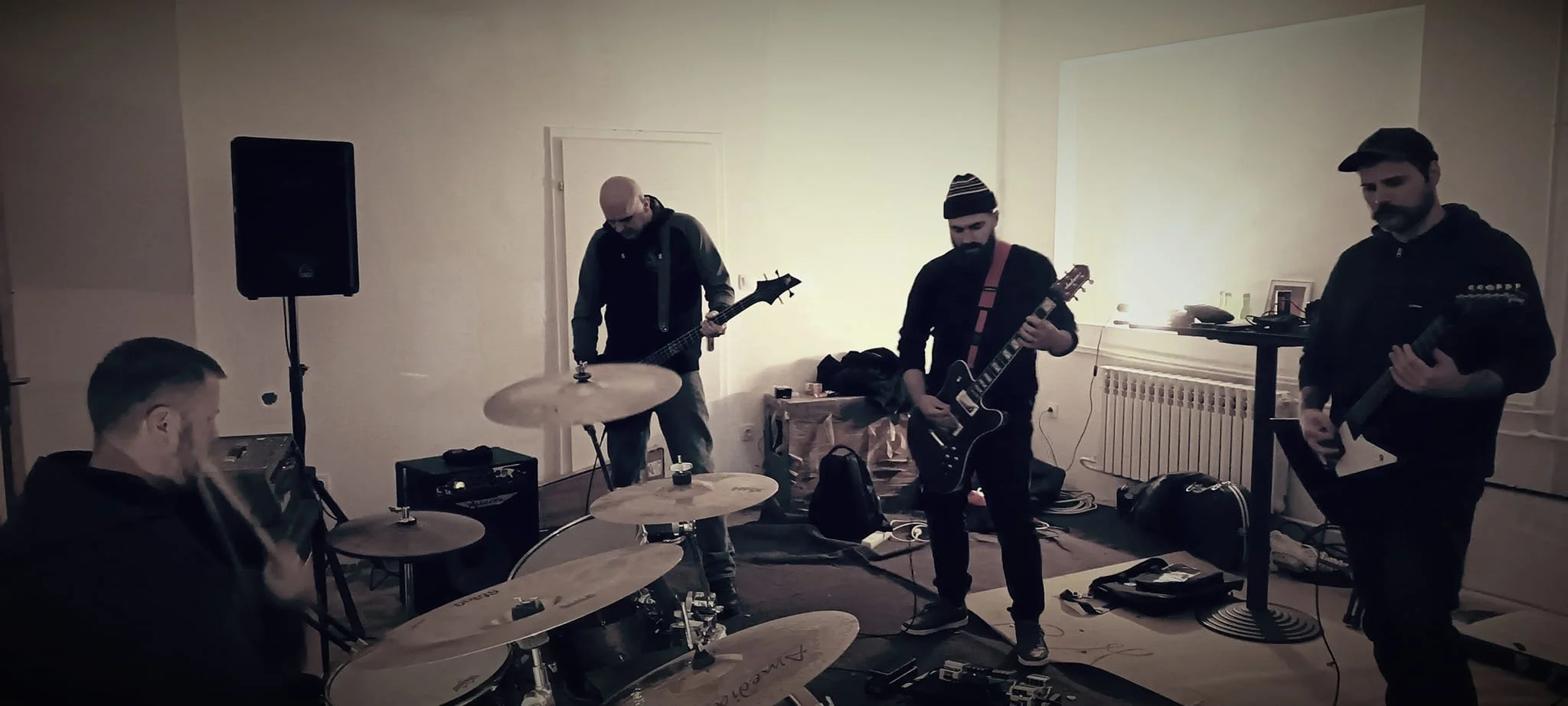 A band of four men playing musical instruments in a room with minimal decor, including drums, guitars, and amplifiers.