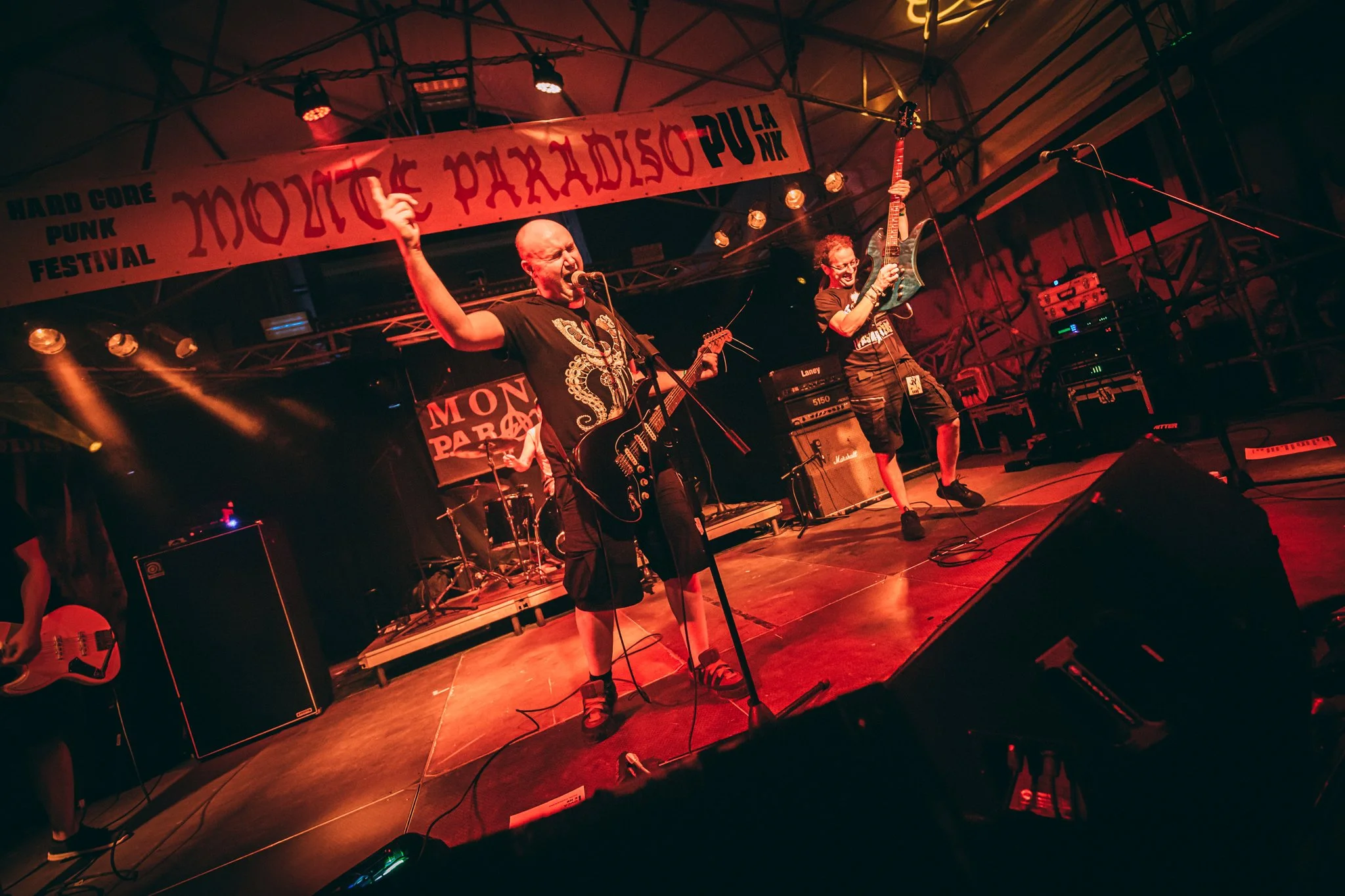 A band performing on stage at a concert with a red lighting ambiance, featuring two musicians in the foreground playing guitars and singing into microphones, with musical equipment and a banner in the background that reads 'Moctezuma Parade' and 'Har