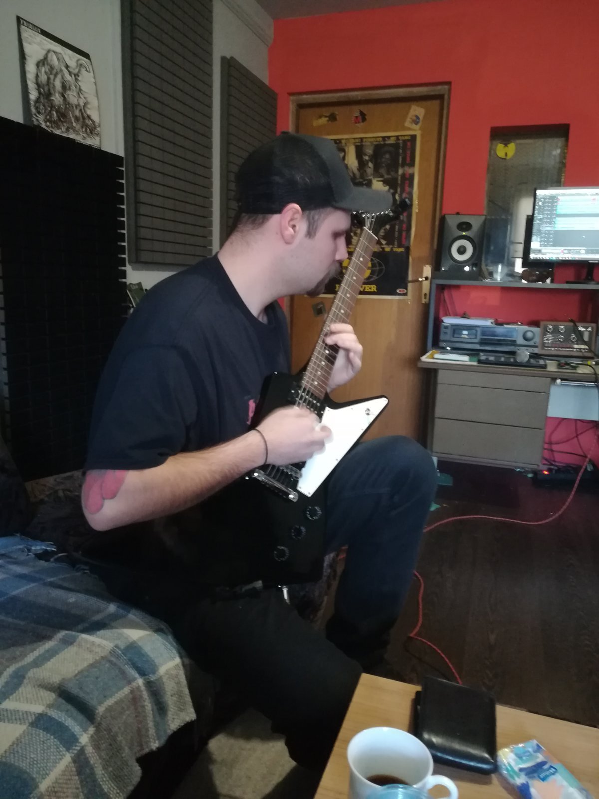 A young man wearing a black cap and black t-shirt is sitting in a music studio, playing an electric guitar. The room has red walls, acoustic foam panels, a desk with audio equipment, a computer monitor, and a speaker in the background.