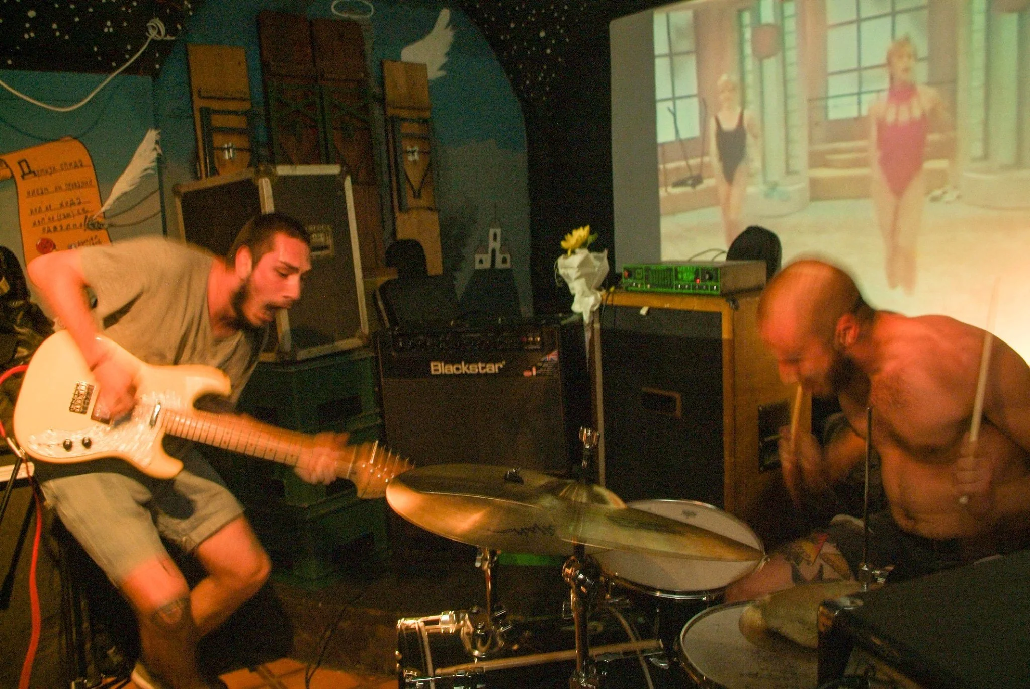 A band performing live in a dimly lit room, with a guitarist playing and a drummer at a drum set. A large screen in the background displays an image of two women in swimsuits.