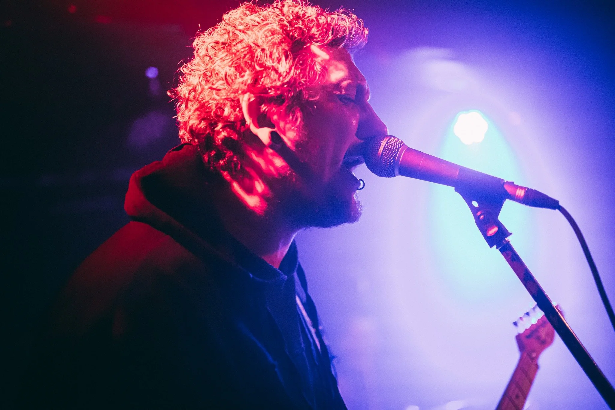A musician with curly hair singing into a microphone on stage, with purple and blue stage lighting.