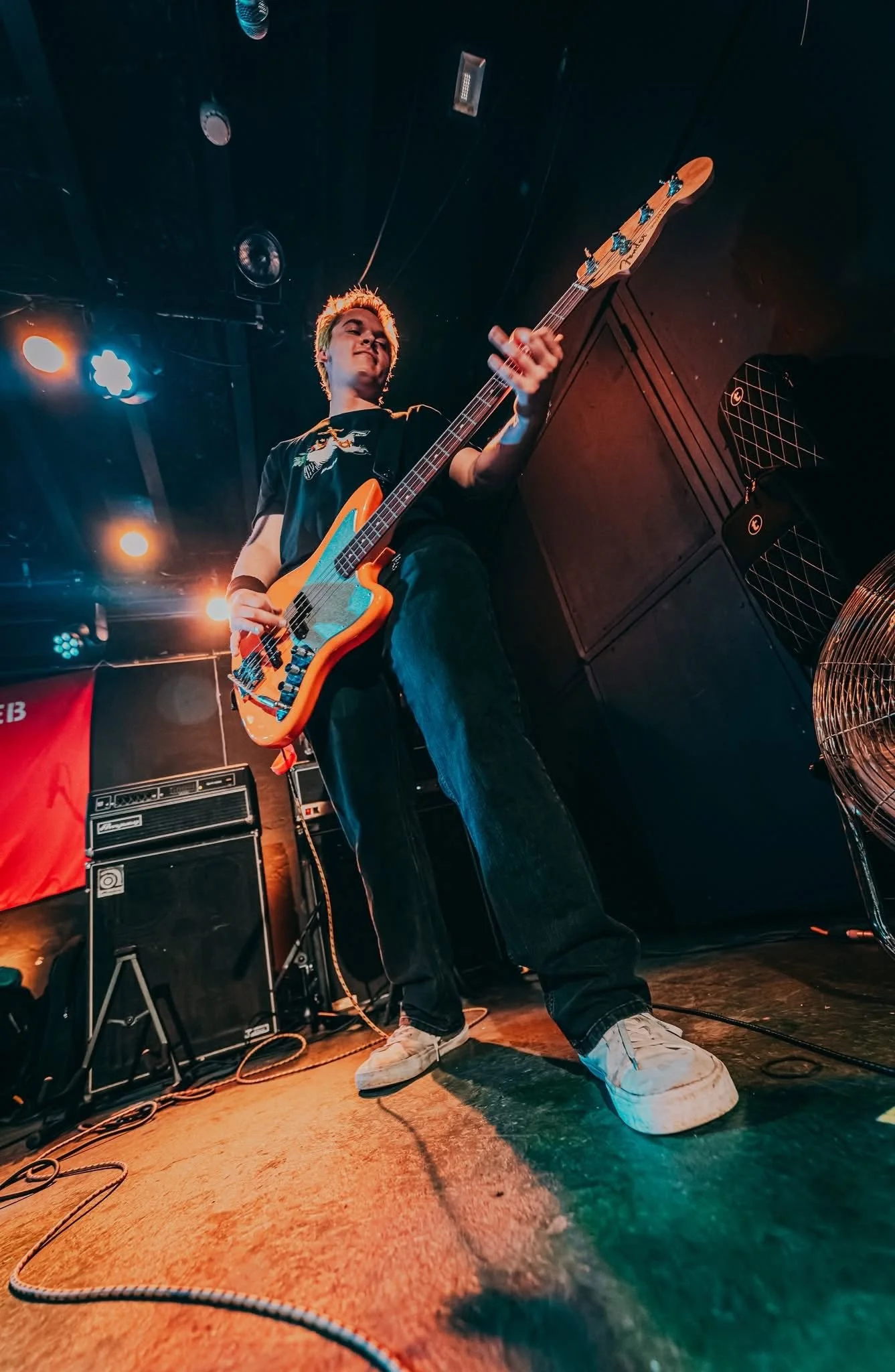 A young musician playing a bass guitar on stage, wearing a black t-shirt and white sneakers, with stage lights and musical equipment visible in the background.