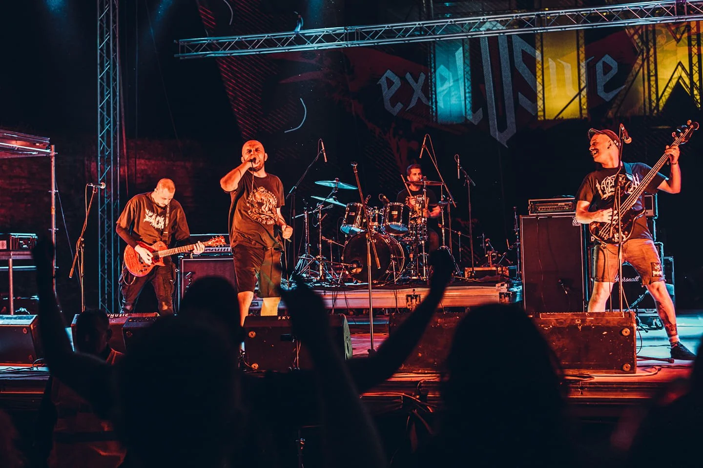 A band performing on stage with four members: a guitarist on the left, a vocalist in the center, a drummer behind, and a bassist on the right. The stage is lit with colorful lights, and audience members are visible with raised hands in the foreground
