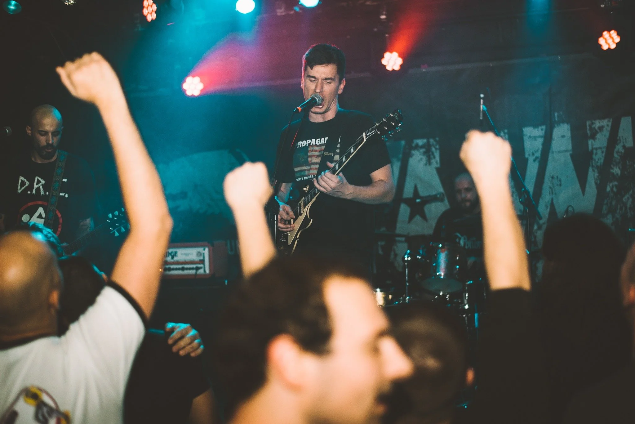 A band performing on stage with a lead singer playing an electric guitar and singing into a microphone, surrounded by colorful stage lights, with audience members raising their fists.