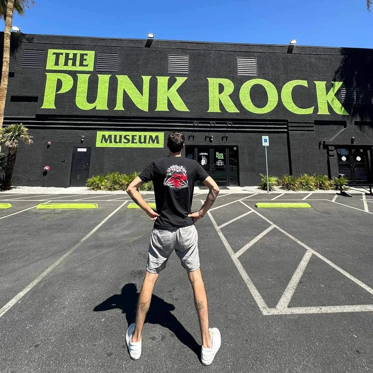 Person standing with hands on hips facing the Punk Rock Museum building with large neon green sign on black wall.
