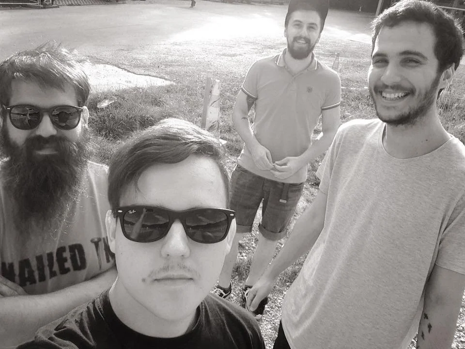 Group of four young men posing outside on a sunny day, with two wearing sunglasses, smiling and standing on a grassy area.