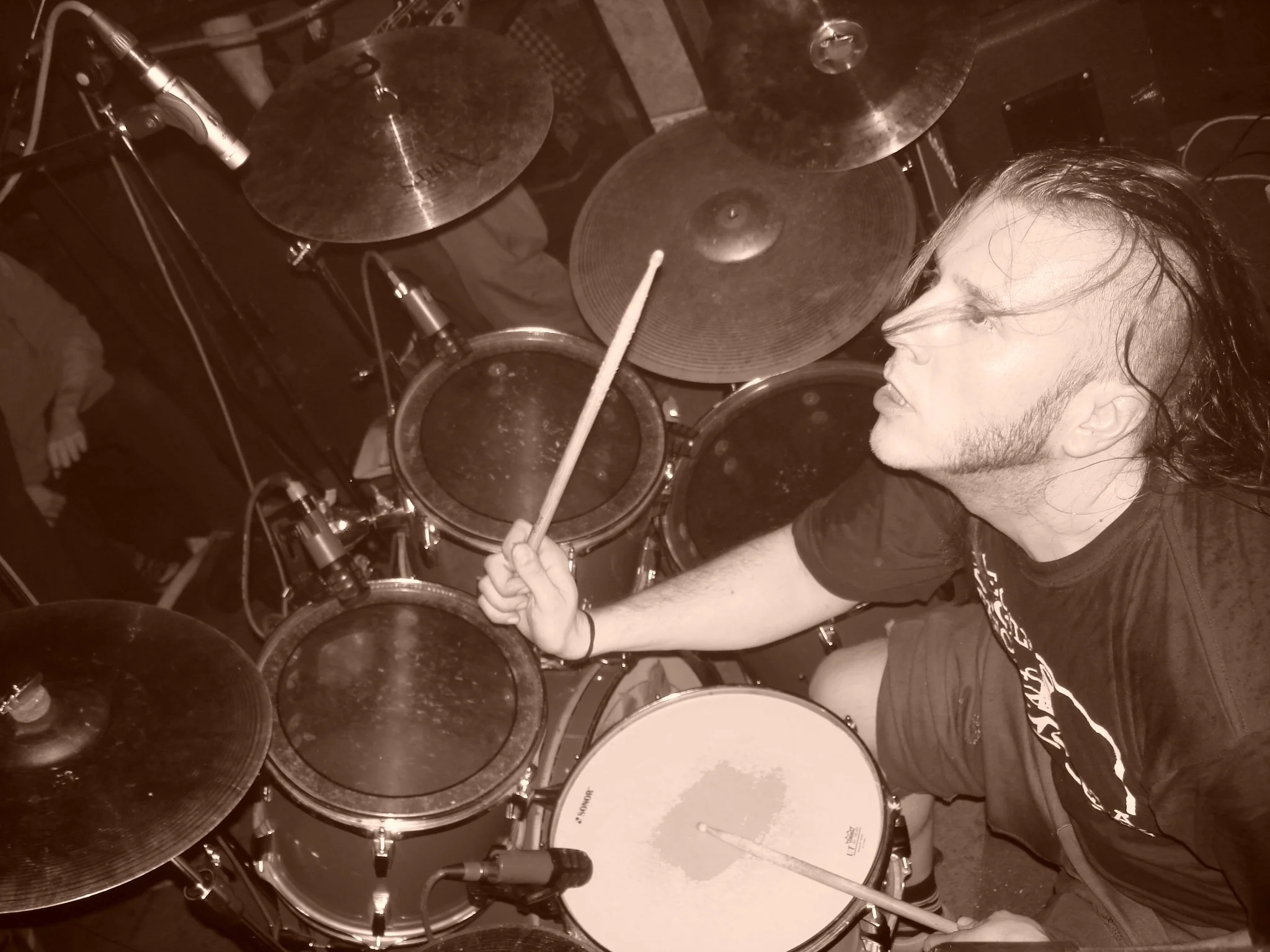 A person with long hair and glasses playing a drum set in a dimly lit room, holding a drumstick in hand near a snare drum. The drum set includes cymbals and various drums, and a microphone is positioned above the set.