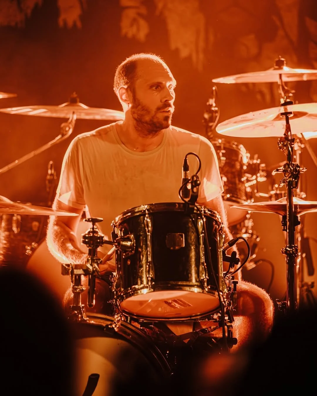 A drummer playing a drum set on stage with orange stage lighting, wearing a wet white t-shirt, surrounded by cymbals.