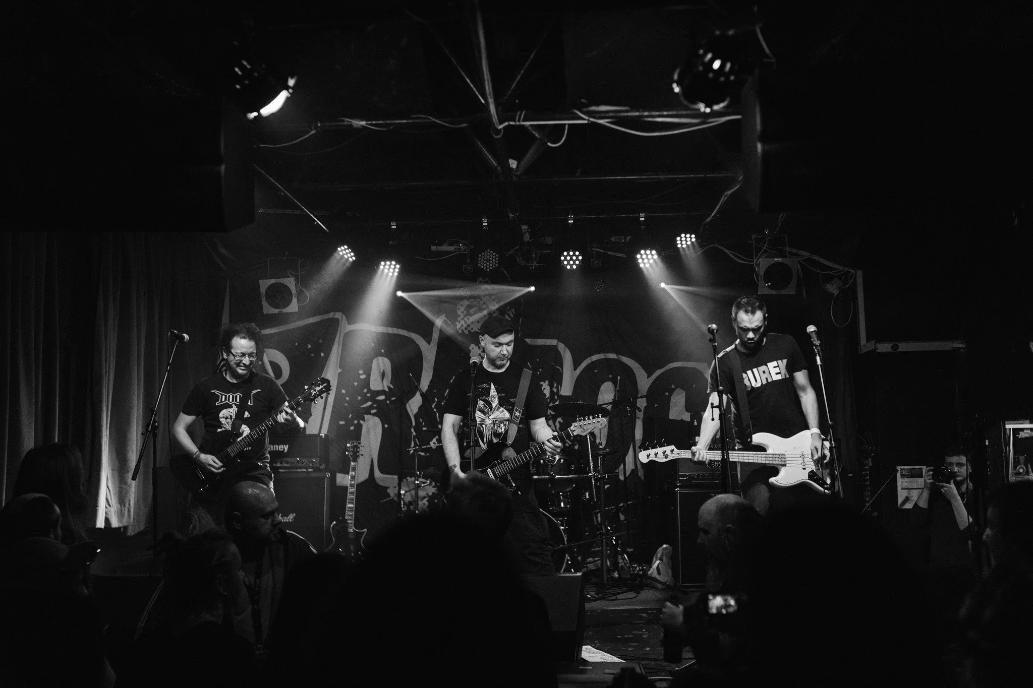 A black and white photo of a band performing on stage, consisting of three musicians playing guitars and a fourth person on keyboard. Audience members are visible in the foreground.