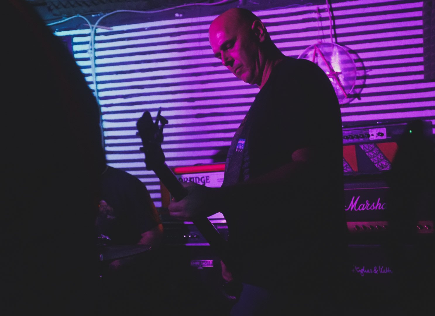 A person playing a guitar in a dimly lit room with neon purple and pink lighting, with musical equipment and a neon sign in the background.